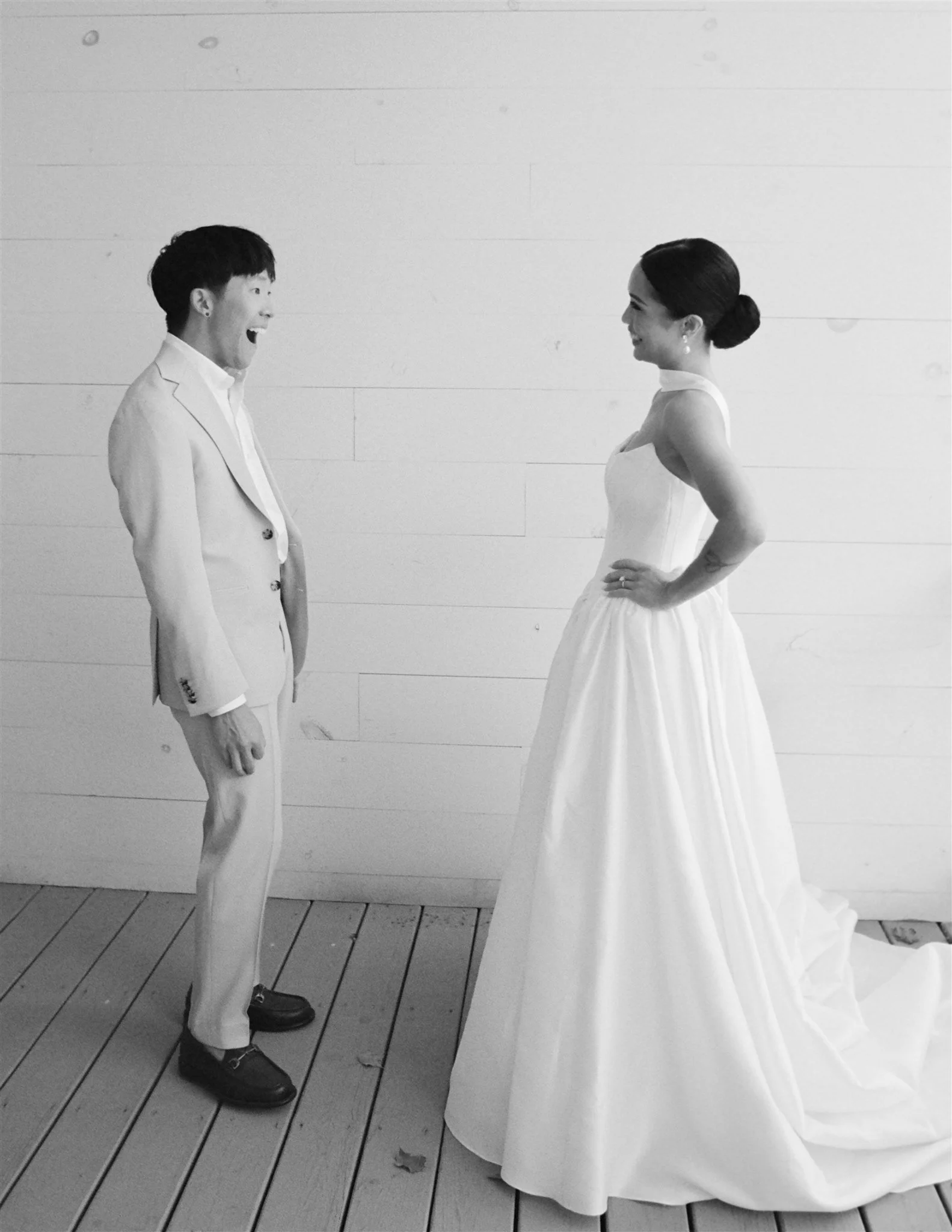Two people face each other, standing indoors against a white wall. The person on the left is a young man with short dark hair wearing a light-colored suit and dark shoes, smiling with an open mouth. The person on the right is a young woman with dark 