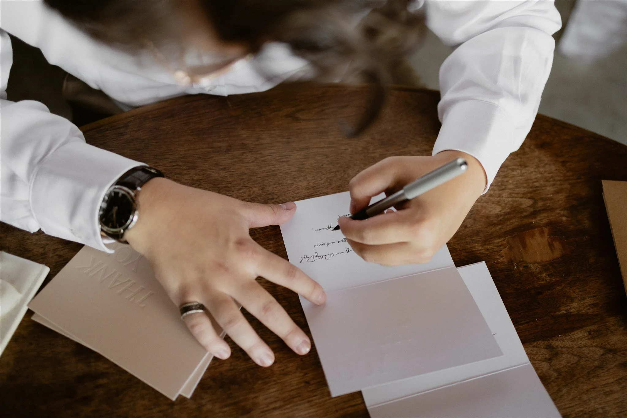 A person with long hair, wearing a white shirt and a watch, is signing a document on a wooden table with a pen. There are papers and a book nearby.