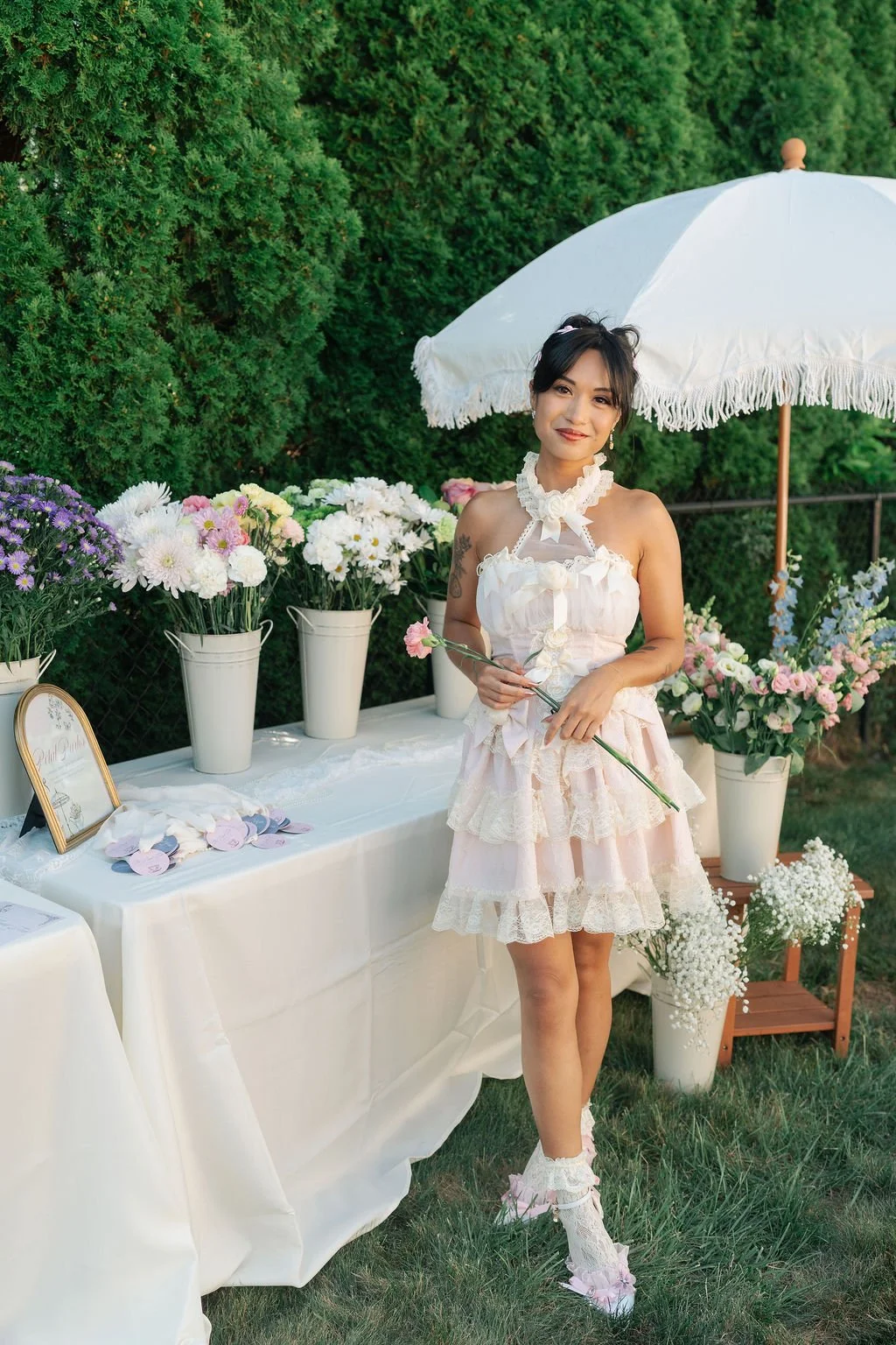 A young woman in a pink and white lace dress holding a pink flower in front of a table with white vases of colorful flowers, standing under a white parasol in a garden setting.