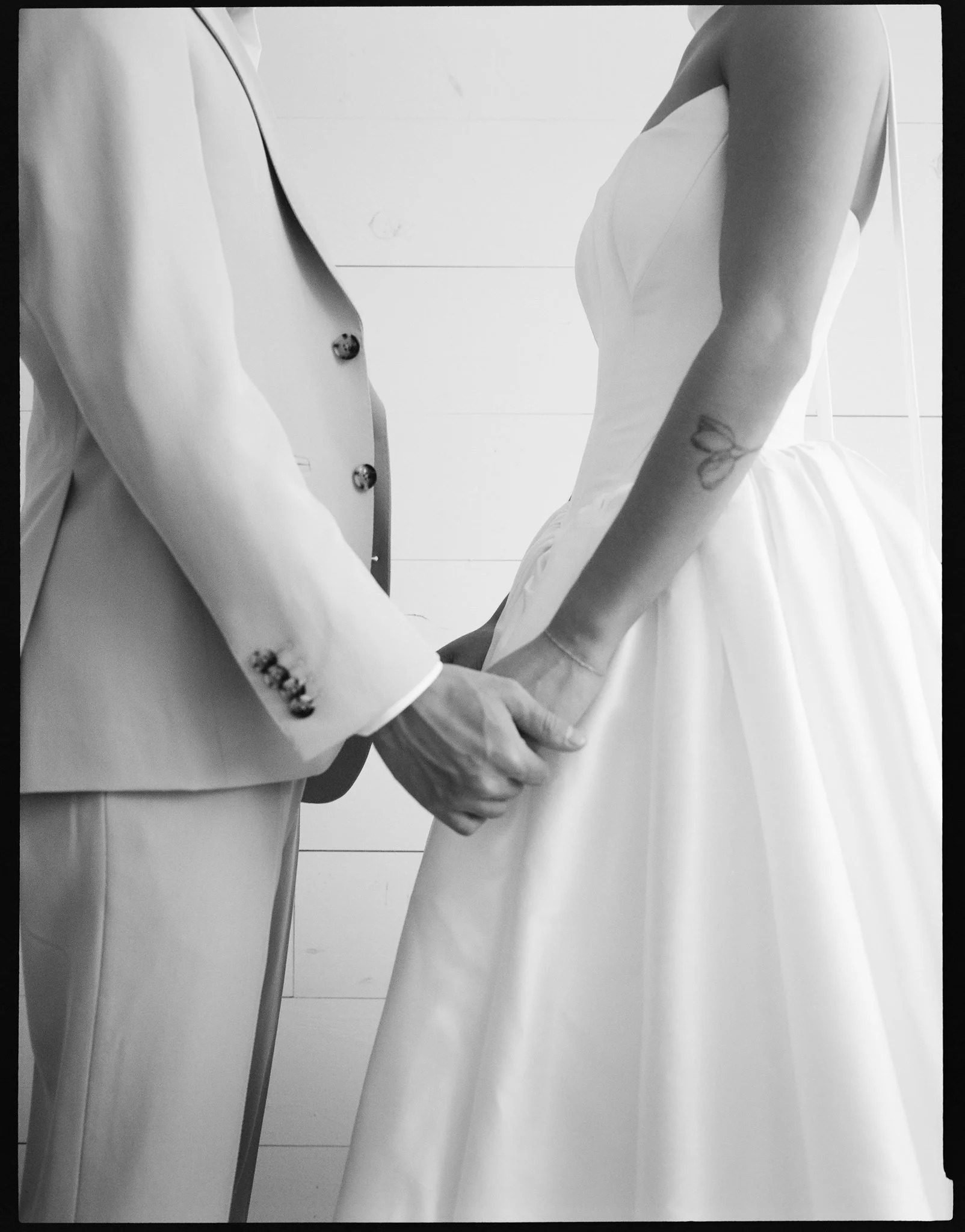Close-up of a couple holding hands in formal attire, wedding dress and suit, on a white tiled background.