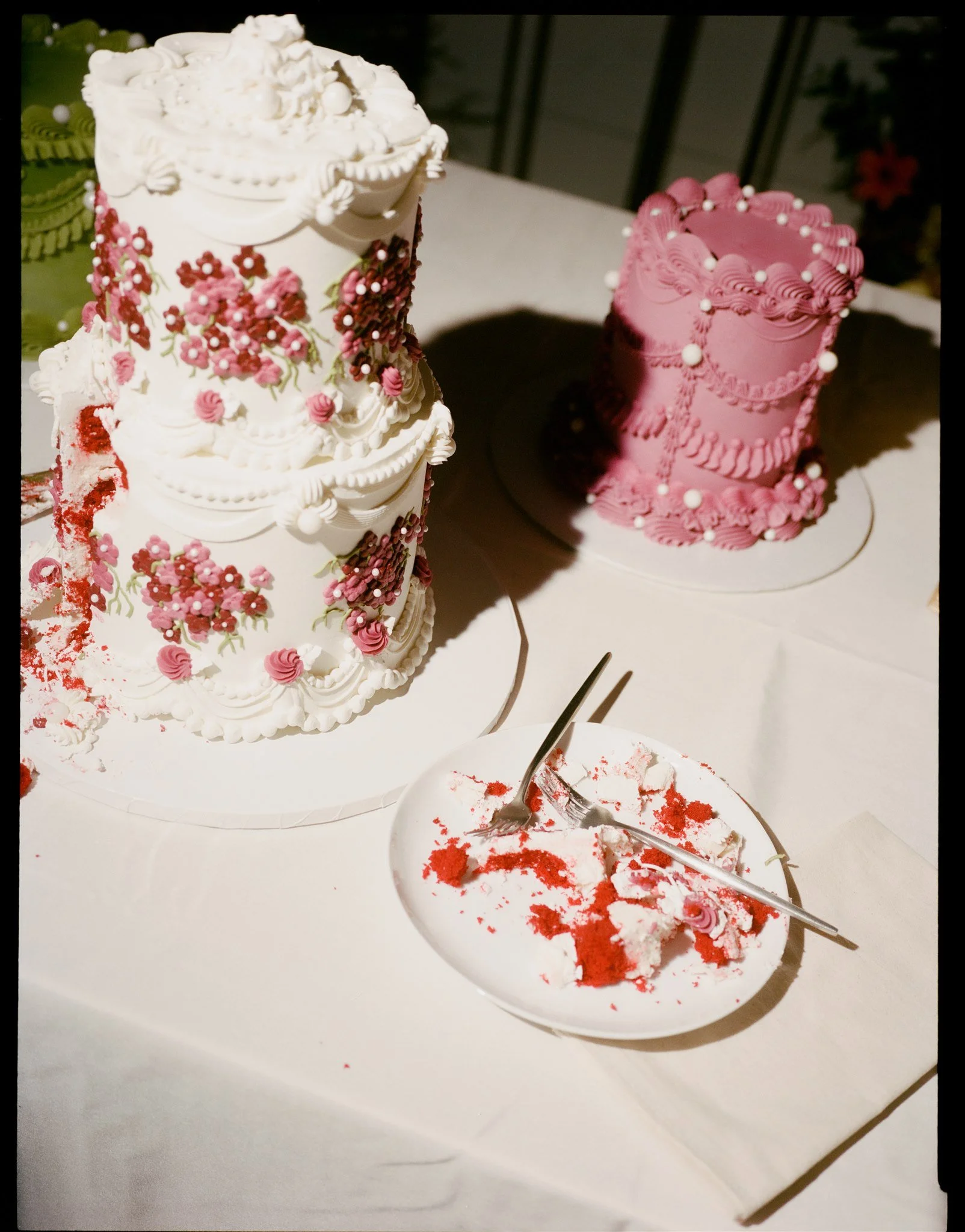 A partially eaten white birthday cake decorated with pink and red flowers and small pearls on top, with a large pink cake with similar decorations in the background, and two forks on a white plate with red cake crumbs and frosting on the table.