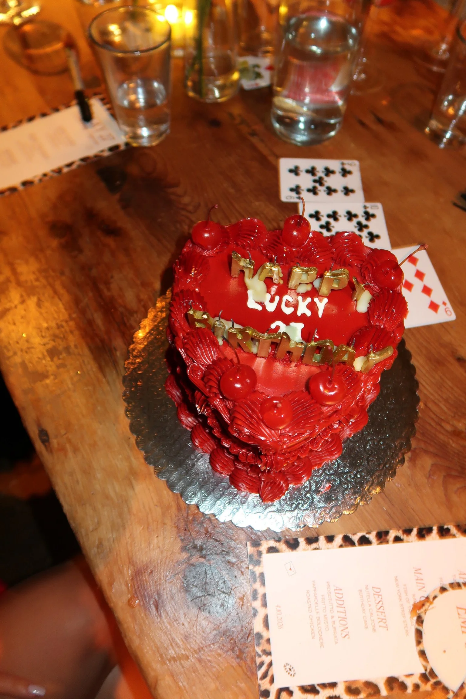 A heart-shaped red birthday cake decorated with cherries and red frosting, with gold and white candles and a birthday message, on a wooden table surrounded by glasses of water and playing cards.