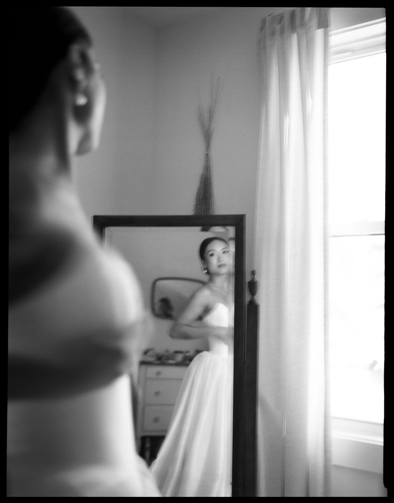 A black and white photo of a young woman looking at her reflection in a mirror, with curtains and a window in the background.
