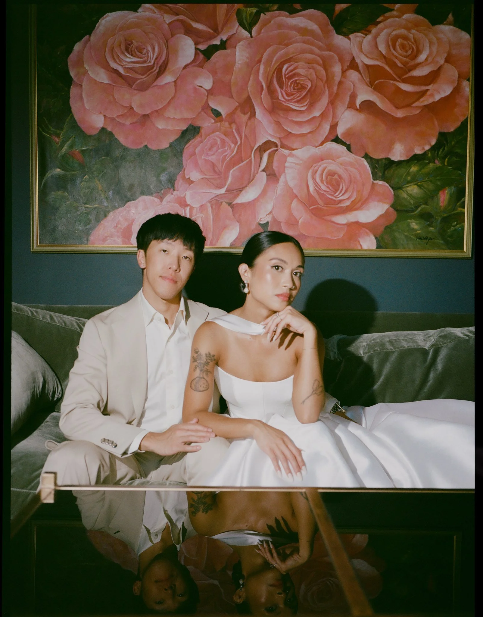 A couple dressed elegantly, sitting on a black leather couch in front of a large painting of pink roses. The man wears a cream suit; the woman wears a white dress with tattoos on her arms and large earrings. Their reflection is visible in a glass or 