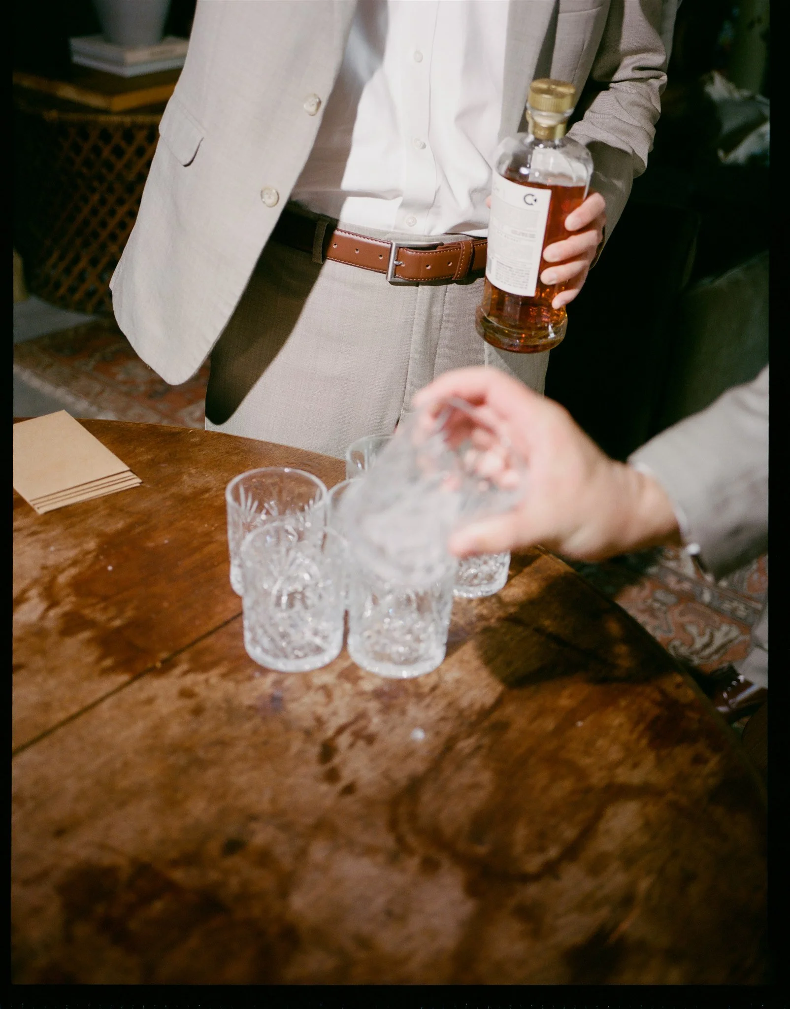 Person in a beige suit and white shirt holding a bottle of liquor, three empty glasses on a wooden table, with a blurred background.
