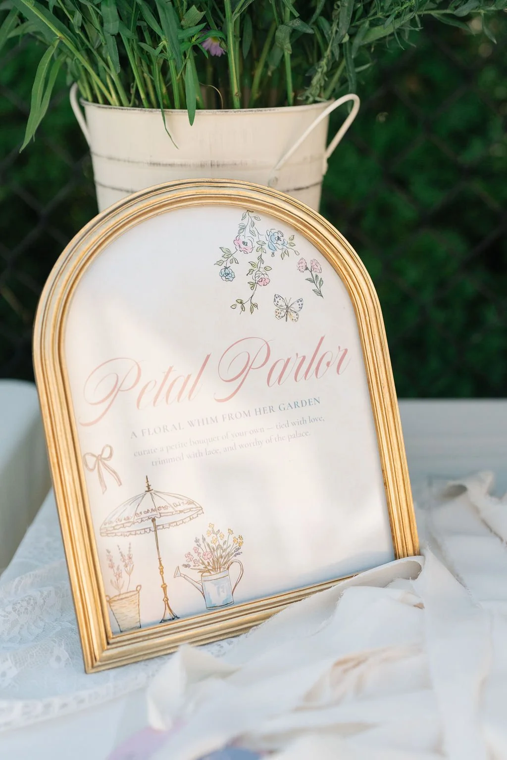 A decorative display featuring a vintage-style sign with floral and garden illustrations, framed in gold, with a white bucket filled with green plants in the background.