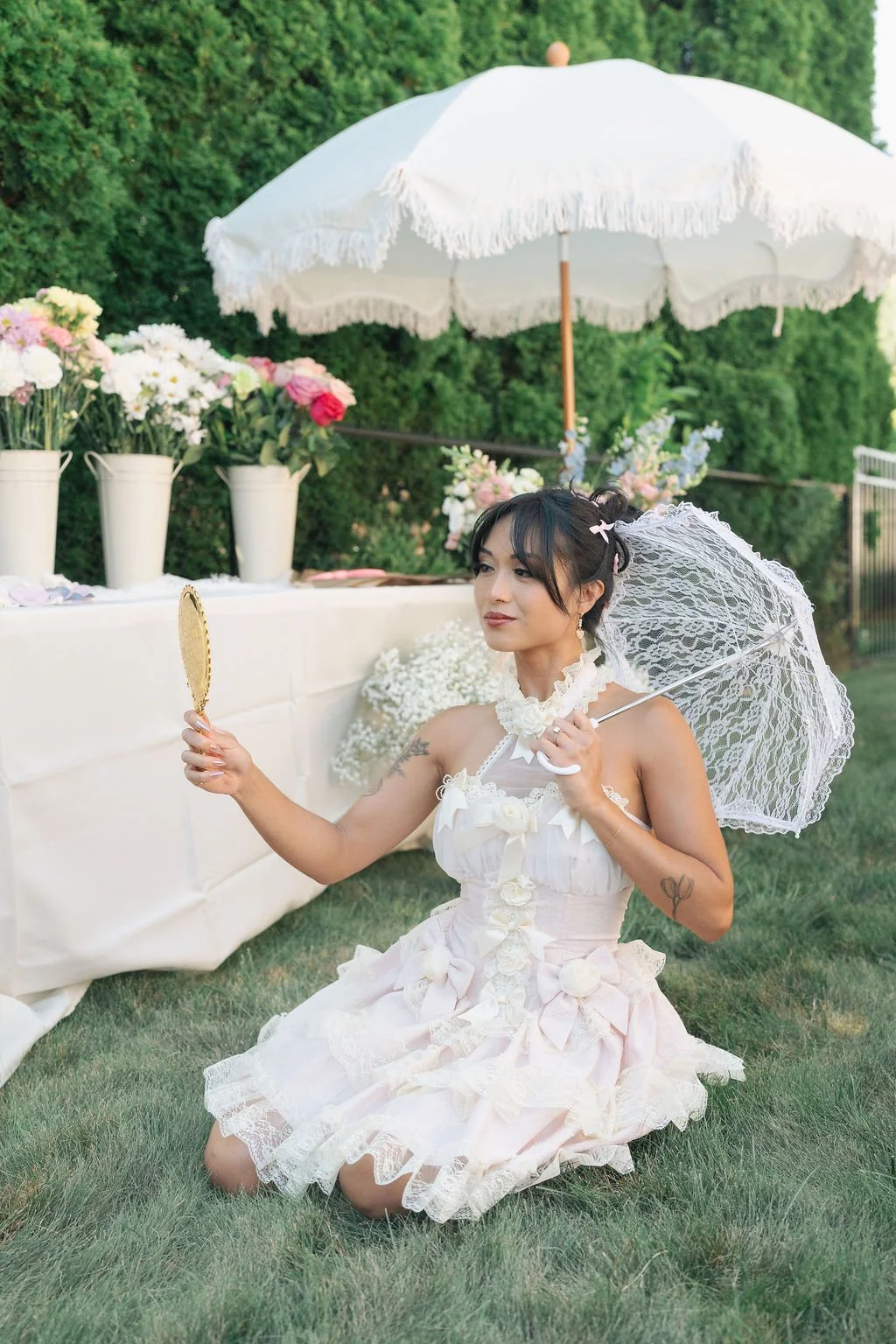 A woman dressed in a white, lace, vintage-style dress kneels on the grass at an outdoor garden party, holding a lace parasol in one hand and a small mirror in the other, surrounded by flower arrangements and a white canopy.