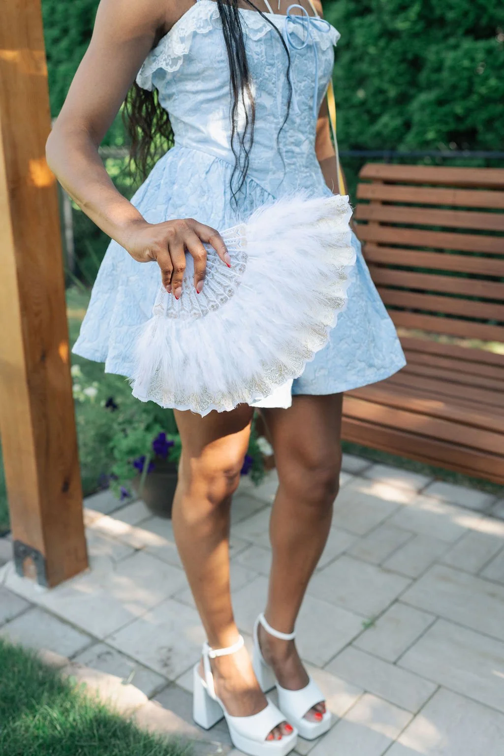 Woman wearing a light blue dress holding a white lace hand fan, standing outdoors on a paved patio with a wooden bench and greenery in the background.