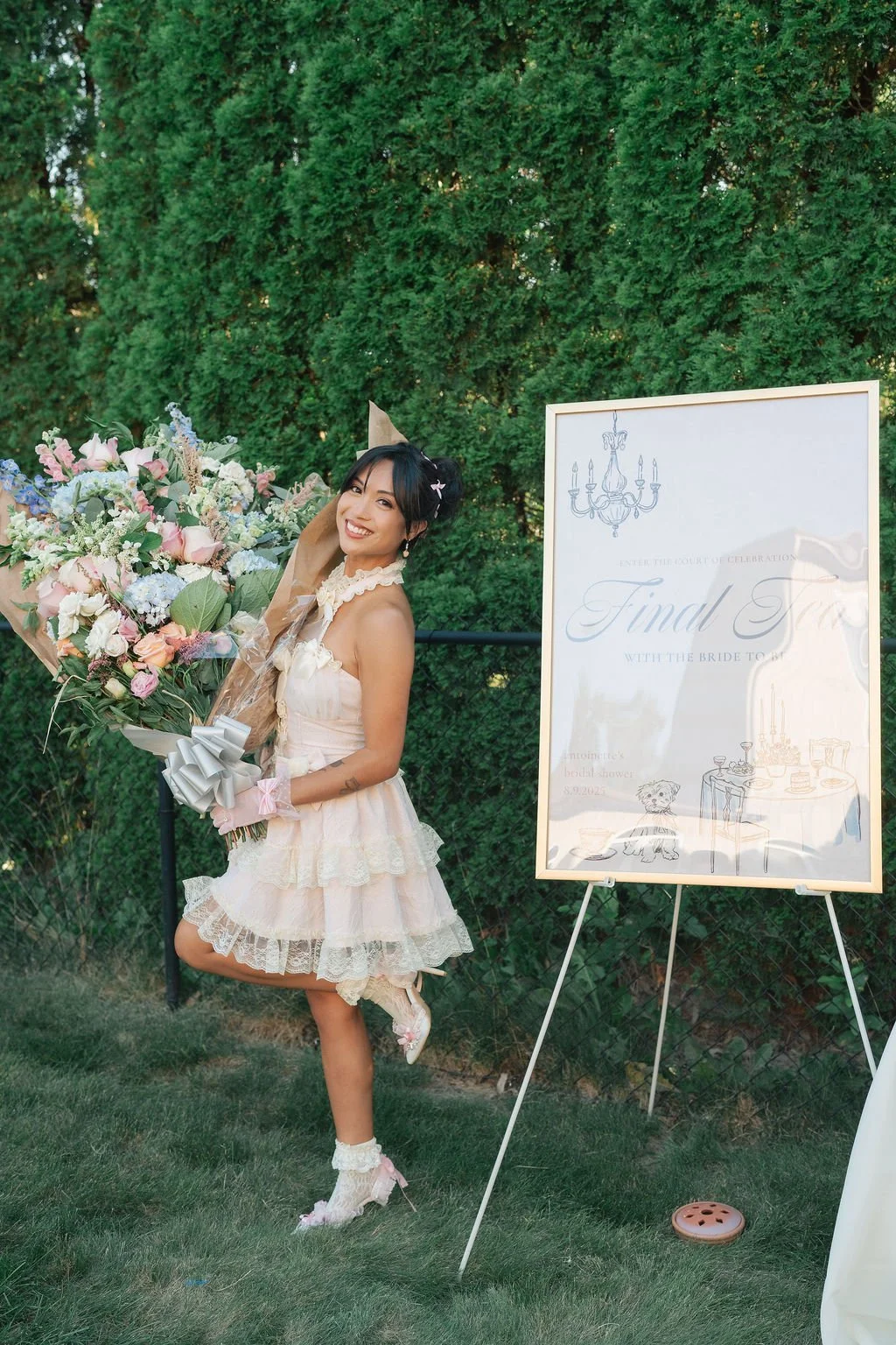 A woman in a light pink, ruffled dress holding a large bouquet of flowers, smiling outdoors next to a sign that reads 'Final Toast' at a celebration event.