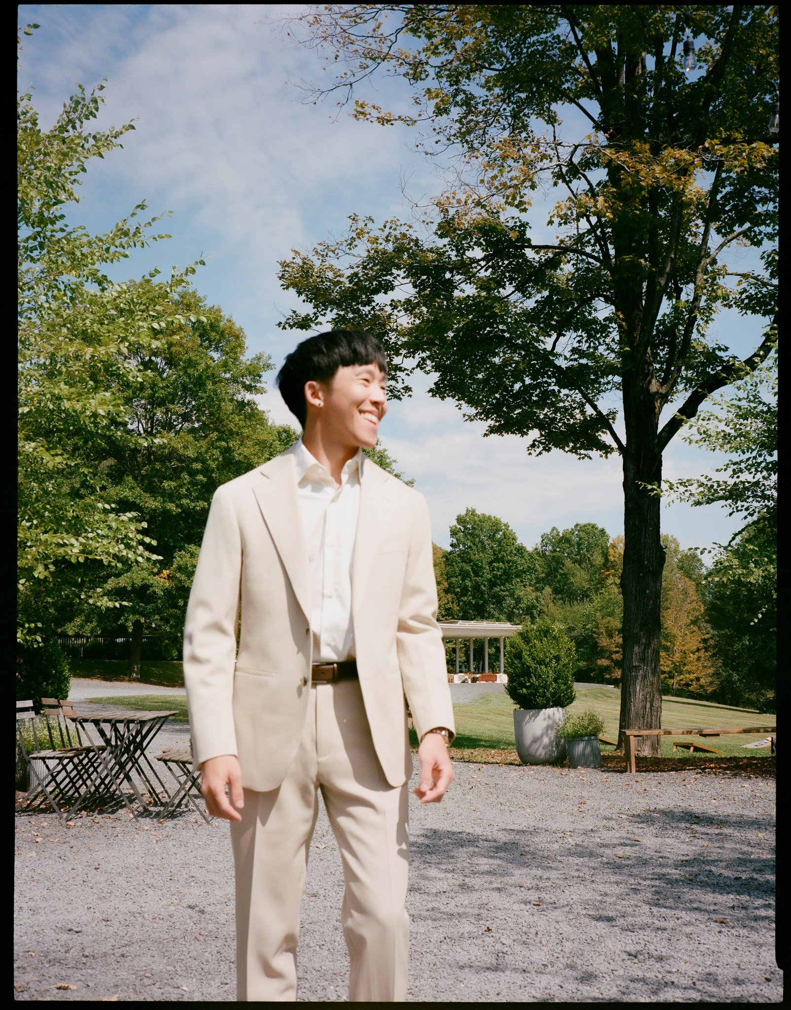 A man dressed in a beige suit outdoors in a park, smiling, with trees, benches, and a gravel pathway visible under a partly cloudy sky.