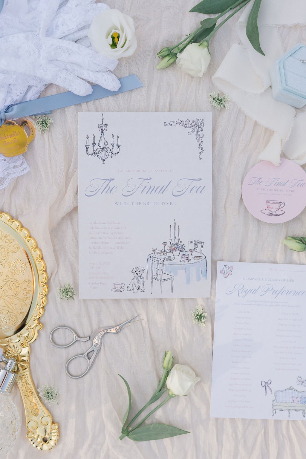 Invitation to the final tea with a chandelier illustration at the top, set on a table with a cake and a teddy bear, surrounded by white flowers, a gold ornate mirror, scissors, and tea-themed decorations.
