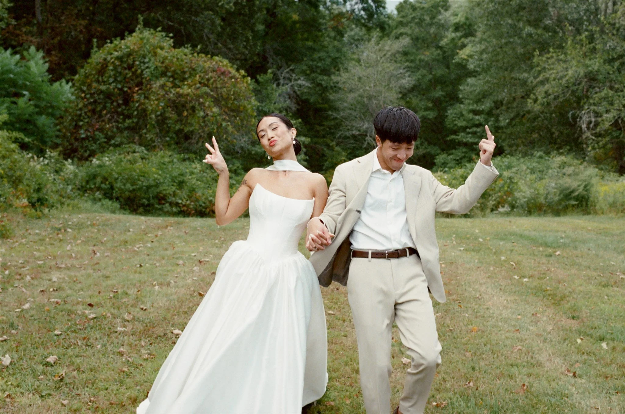 A woman in a white wedding dress and a man in a beige suit holding hands and dancing outdoors in a grassy area surrounded by trees.