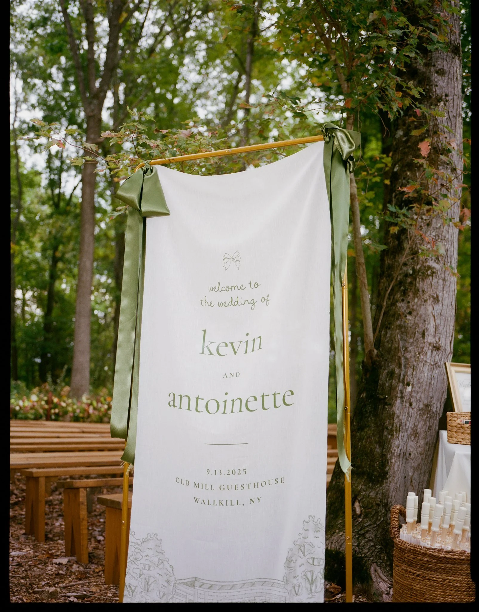 A wedding welcome sign hanging outdoors between trees, with a white background and green ribbon accents. The sign reads, 'welcome to the wedding of kevin and antoinette, 9.13.2025, old mill guesthouse, Wallkill, NY,' with a faint outline of trees and