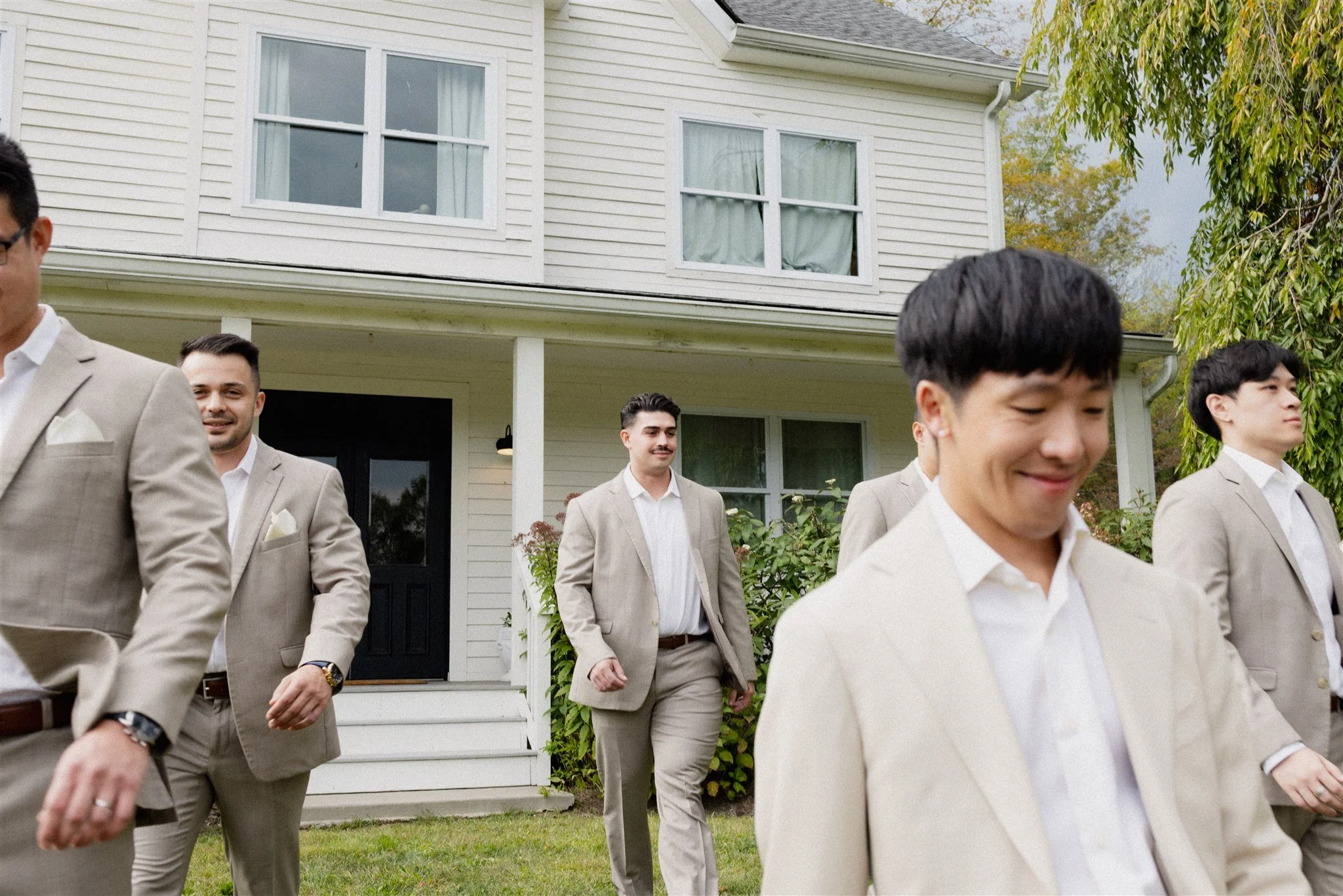 Group of men in beige suits walking outside a white house with large windows and greenery.