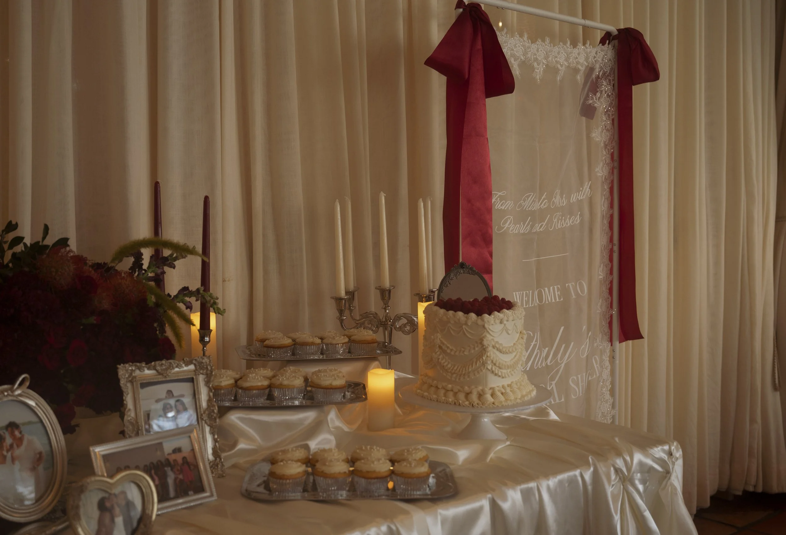 A display table with a wedding cake, cupcakes, framed photographs, and lit candles in a decorated setting with beige curtains and red ribbons.
