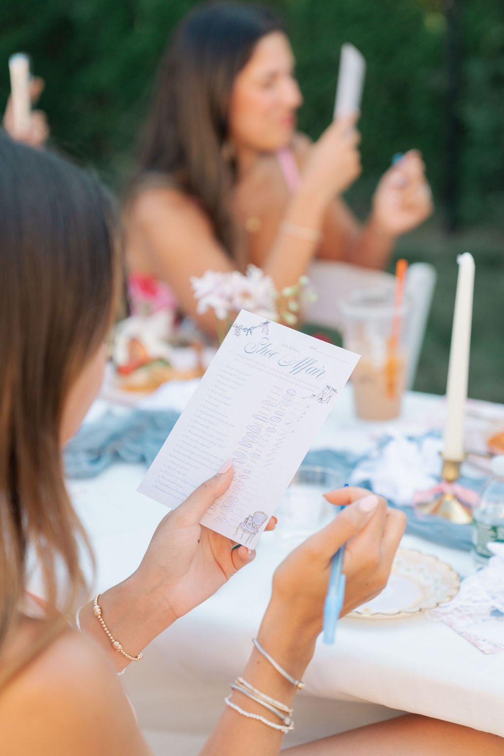 Person at a table reading a wedding program while sitting outdoors at a celebration with candles, drinks, and table decorations.
