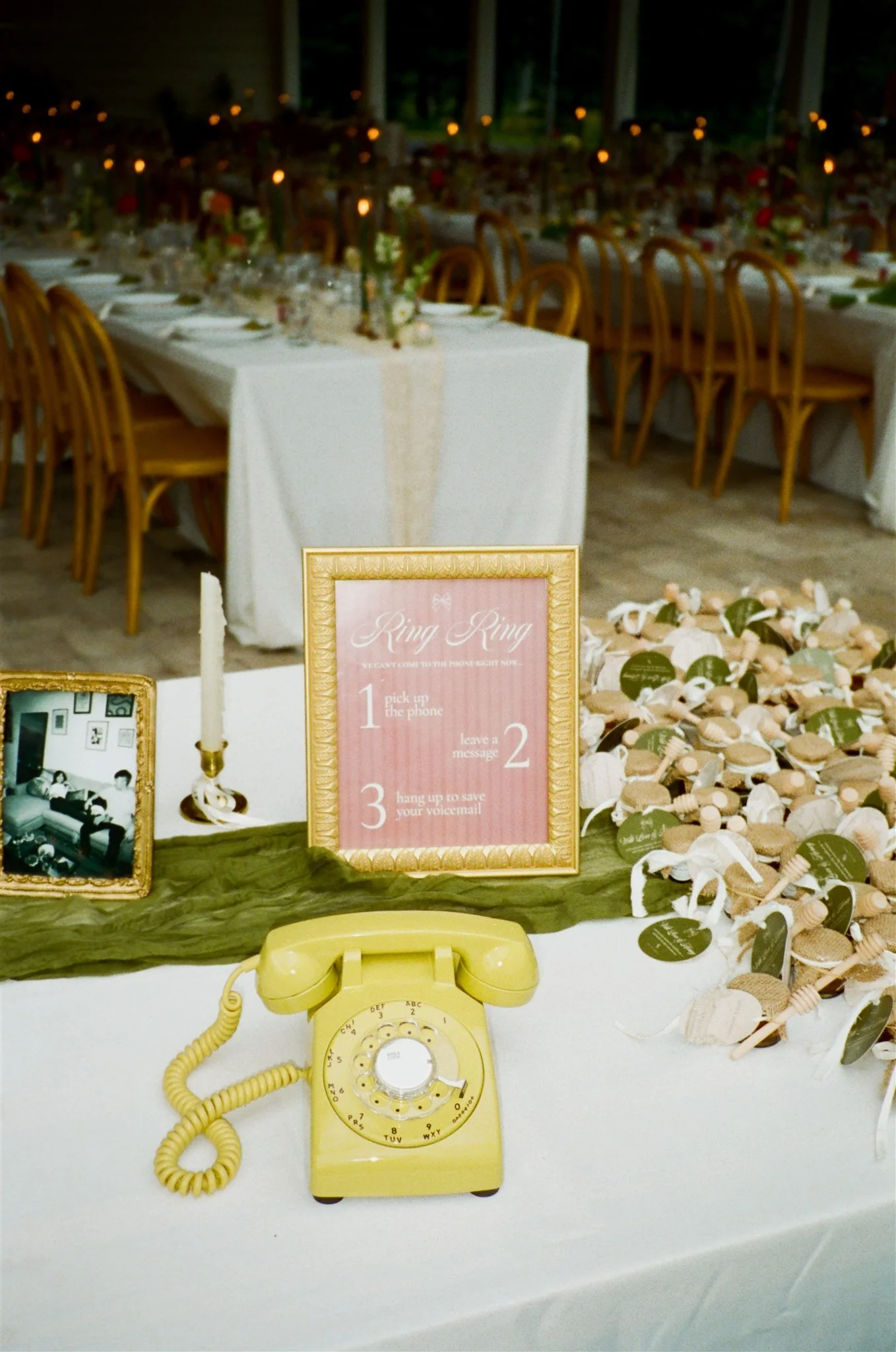 A yellow rotary telephone on a white table, with a wooden tray holding seashells and a pink sign with instructions, in a decorated event space.