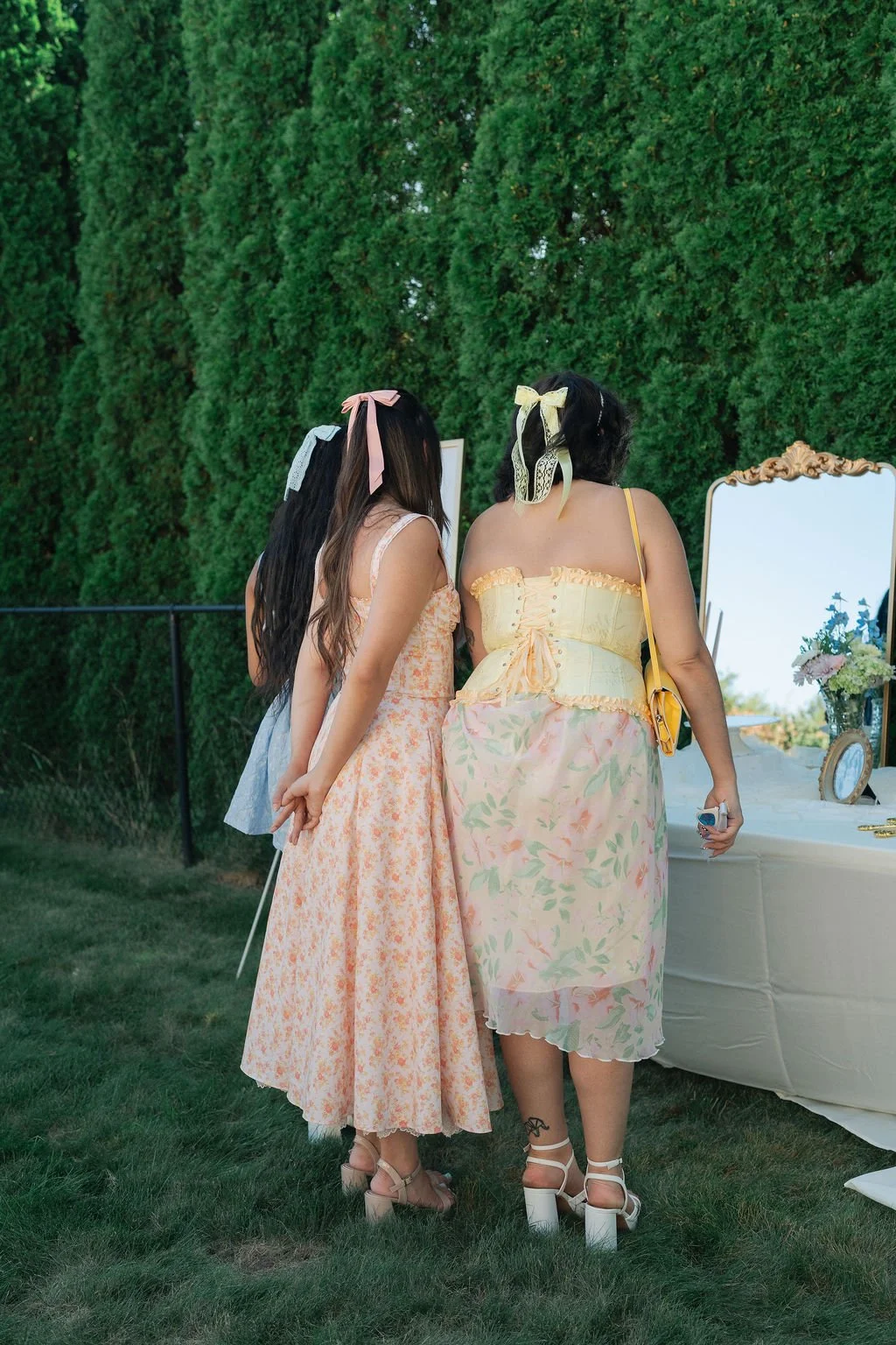 Two women standing outdoors in front of a large mirror and a table with flowers, dressed in pastel-colored dresses and wearing headbands with ribbons.