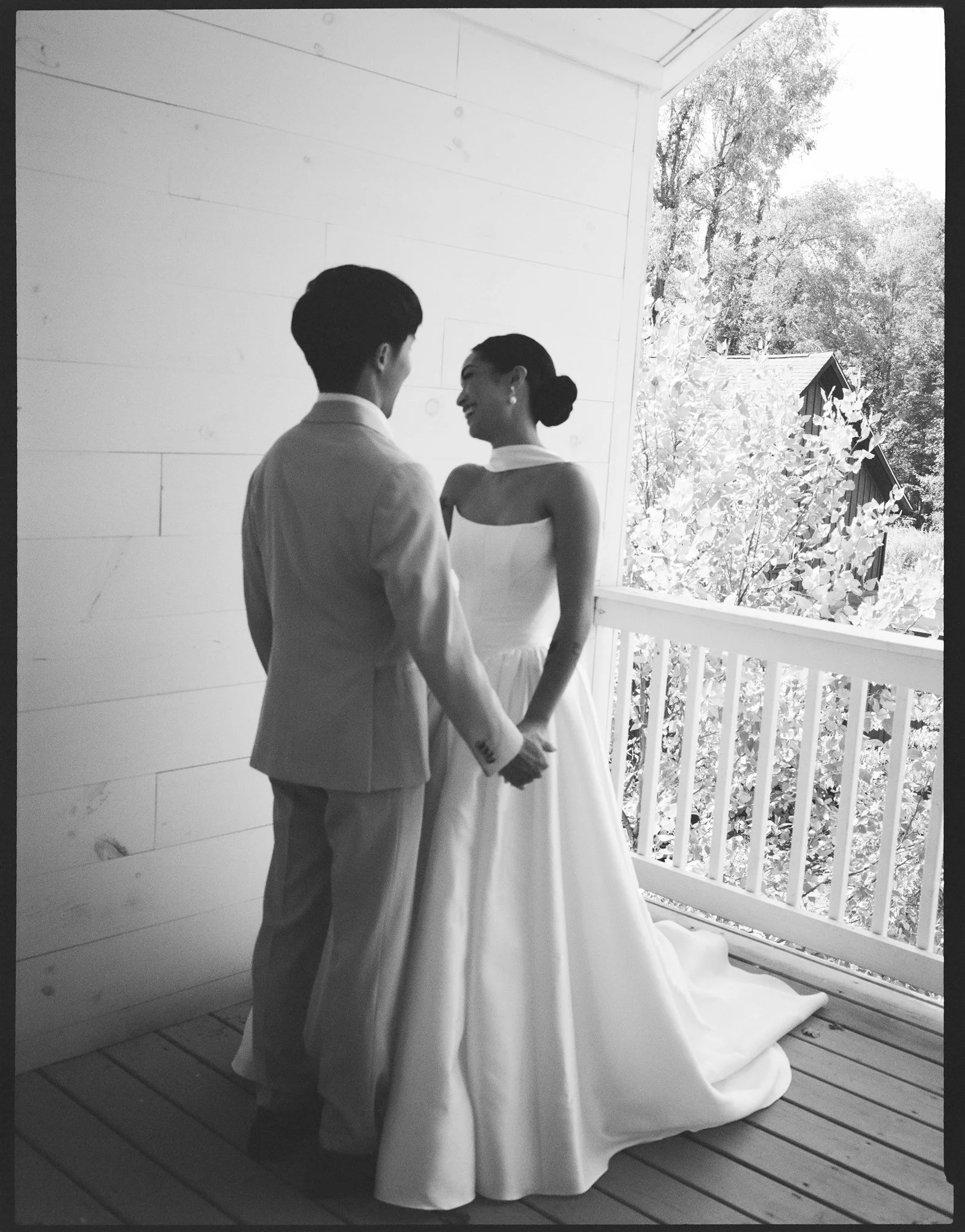 A bride and groom holding hands and looking at each other on a porch with wooden flooring, a white railing, and open windows. The bride is wearing a white wedding dress, and the groom is dressed in a light-colored suit. Outside, trees and a small bui