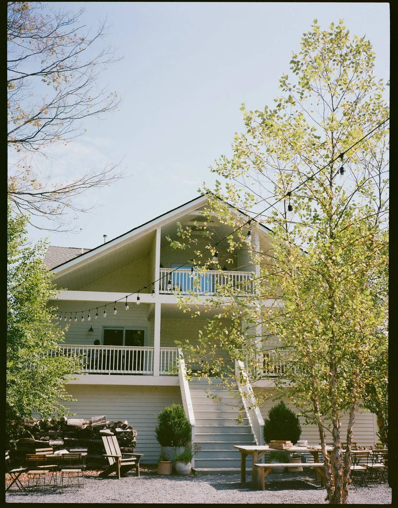 Backyard patio area with wooden furniture, a firewood stack, potted plants, string lights, and a house with stairs leading to a balcony, surrounded by trees with fall foliage.