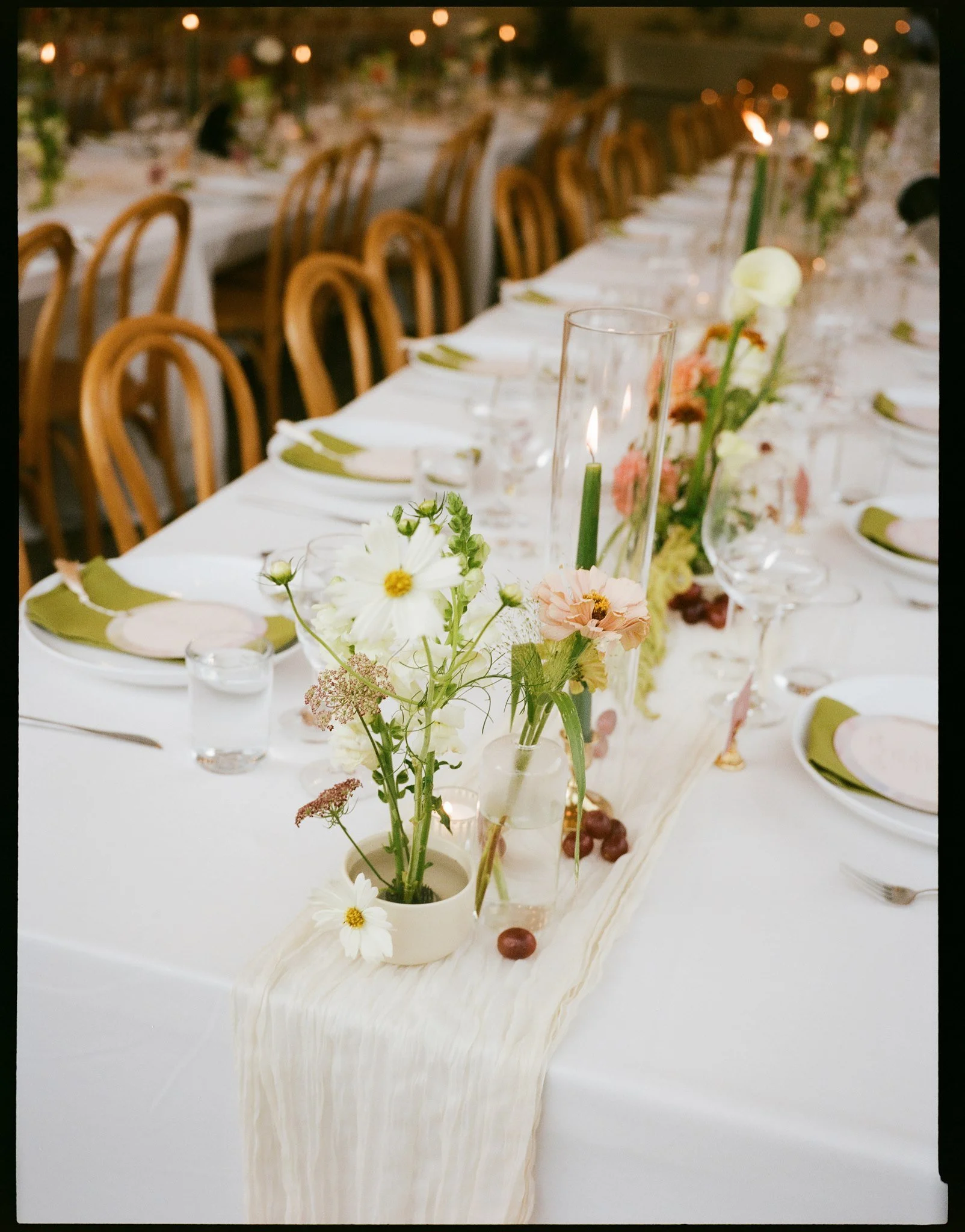 Long wedding reception table decorated with a cream-colored cloth, floral arrangements in small vases, and floating candles, with wooden chairs and blurred string lights in the background.