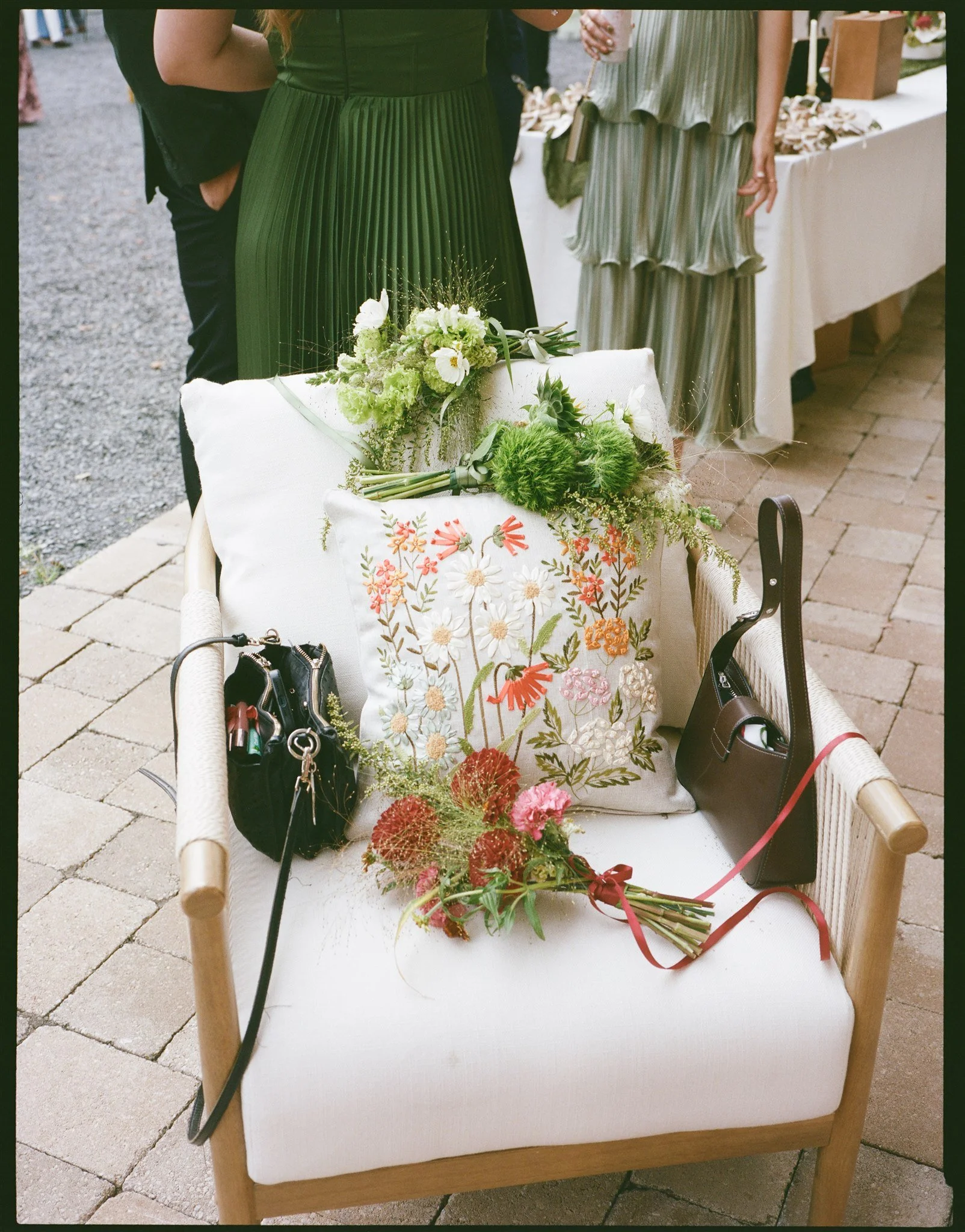 A white cushioned sofa with embroidered floral patterns in orange, pink, and white, decorated with green leaves. On the sofa are a small black purse, a black handbag, and a bouquet of flowers including red, pink, and green blooms. The background show