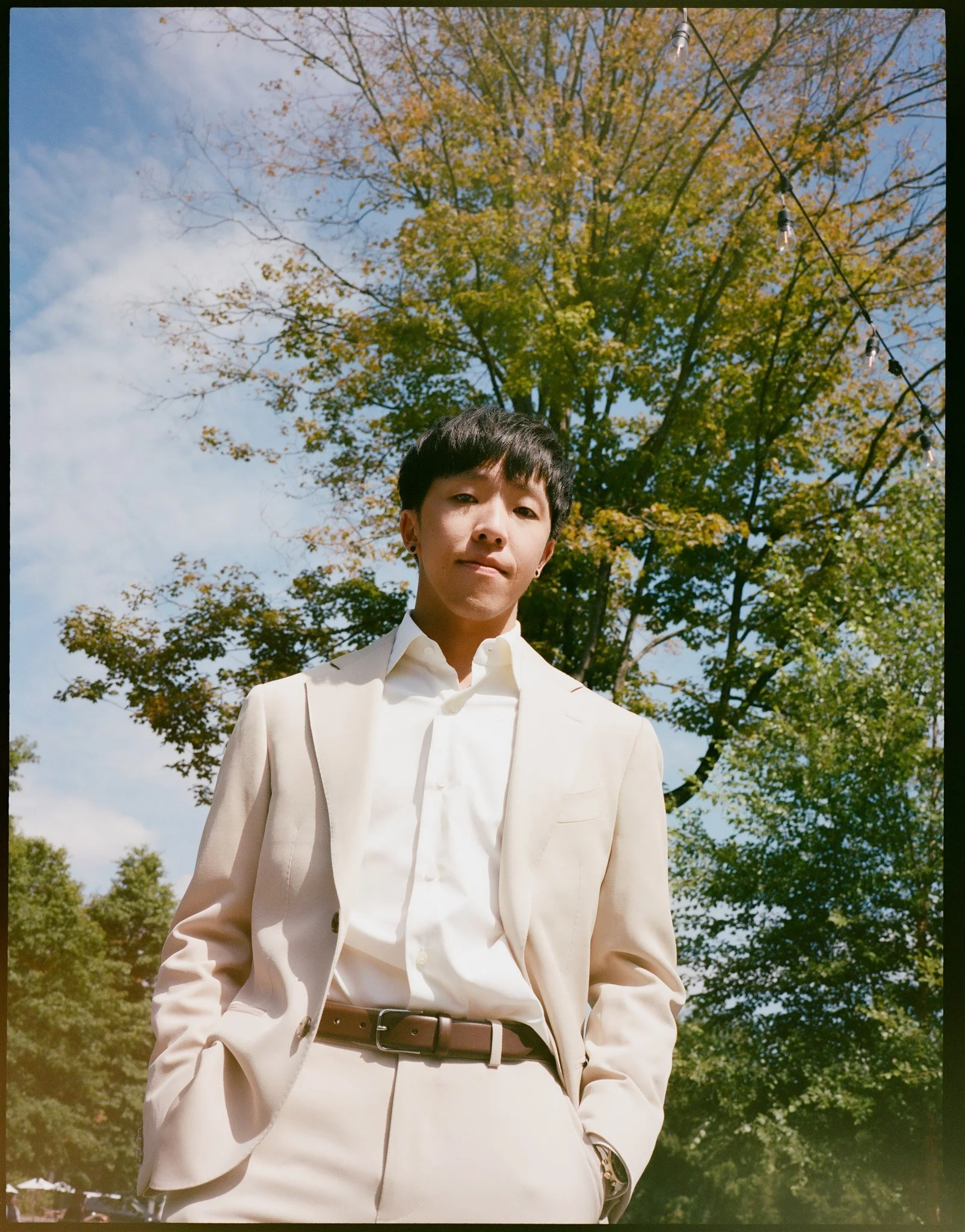 A person with short dark hair and earrings wearing a white suit with a belt, standing outdoors under a large tree with green and yellow leaves on a sunny day, with string lights hanging overhead.
