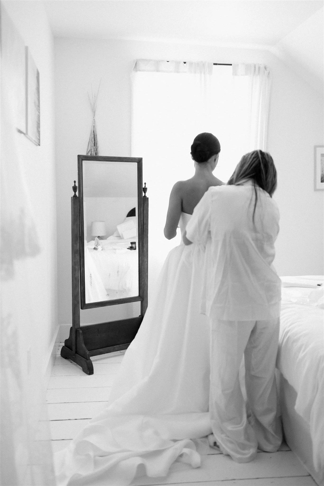 A woman draped in a white sheet kneels on a white floor next to a mirror, with her back to the camera. The room has white walls, and there is a window with white curtains. A bed with white bedding is reflected in the mirror, and a framed picture hang