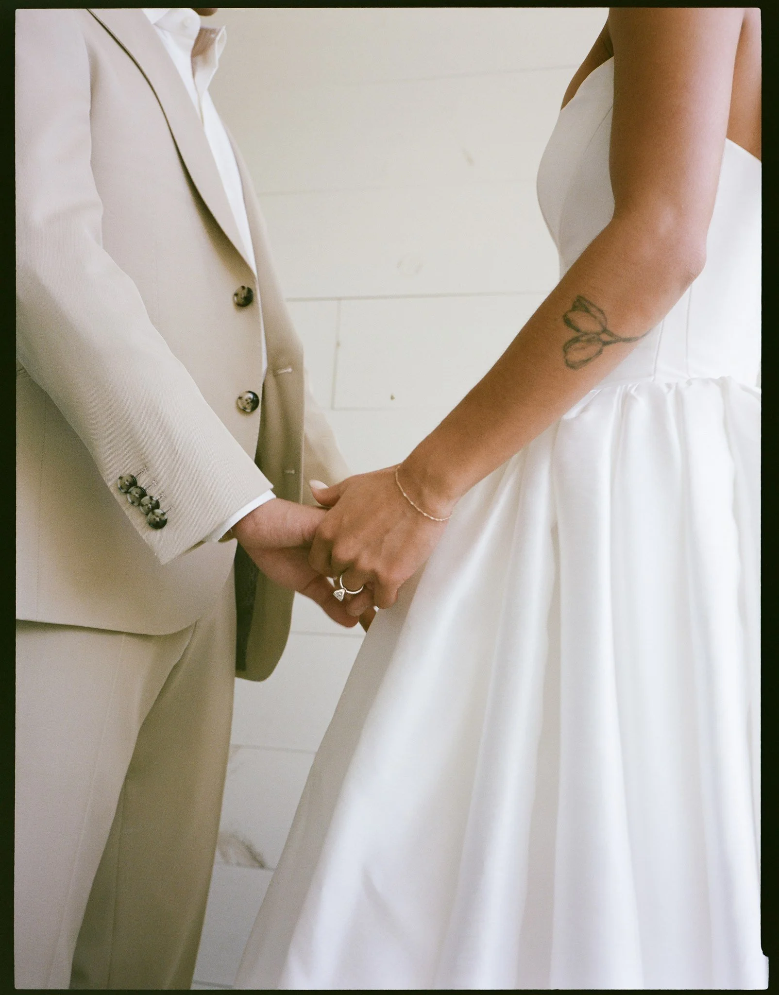 A couple holding hands, one wearing a white suit and the other wearing a white dress with a butterfly tattoo on their arm.