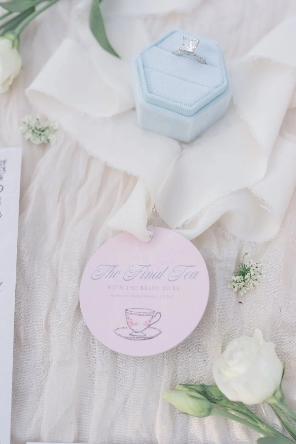 A light blue ring box with a large diamond engagement ring inside, placed on a white fabric surface with flowers and a pink card reading 'The Final Tea with the Bride to Be' featuring a tea cup illustration.