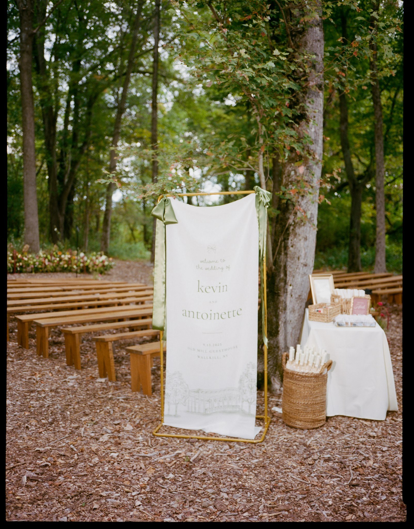 Outdoor wedding setting with wooden benches, a fabric wedding sign with green ribbons, and candles on a table, all surrounded by trees and leaf-covered ground.