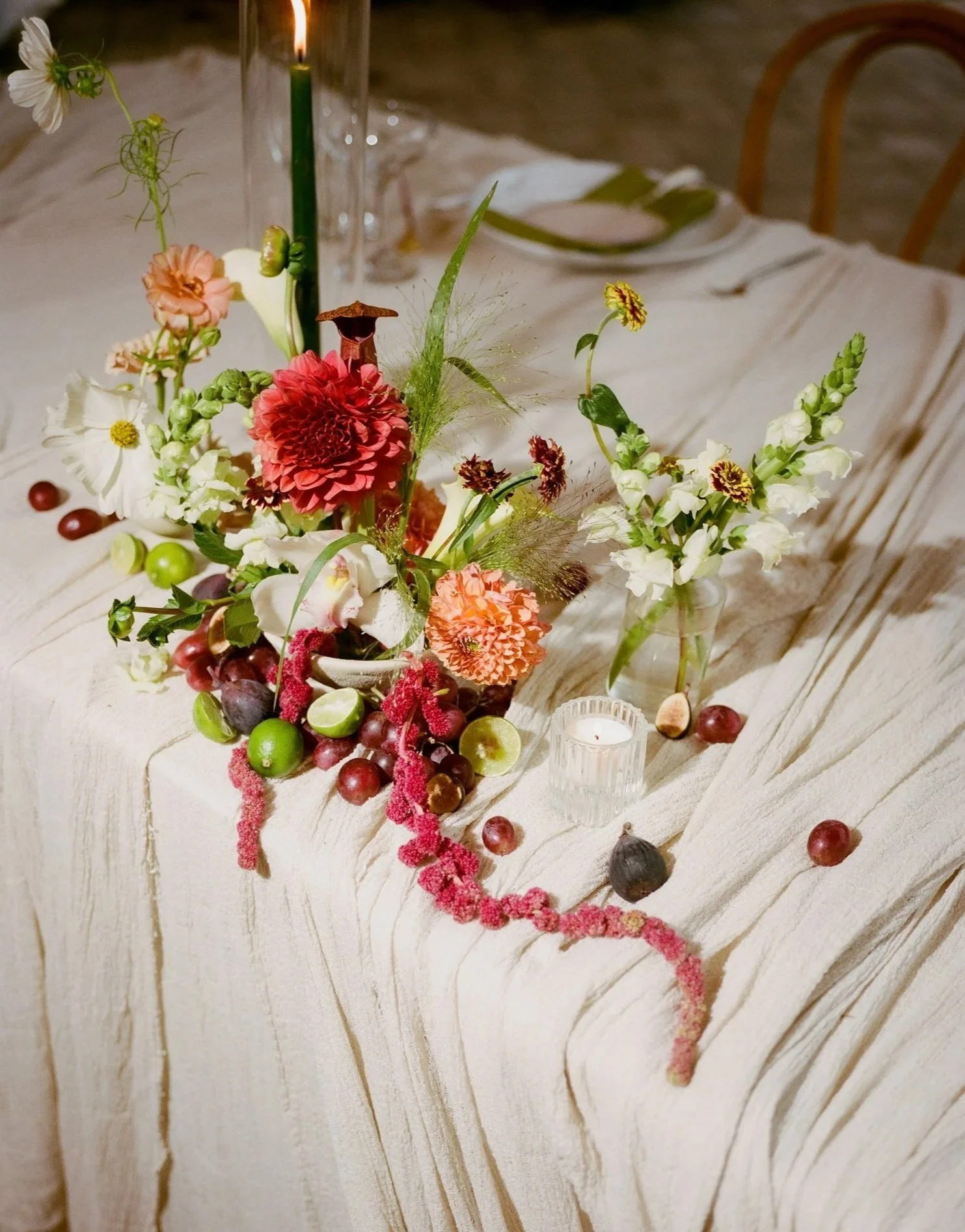 A floral centerpiece with pink, white, and orange flowers, green foliage, a tall green candle, and scattered grapes and figs on a white wooden table.