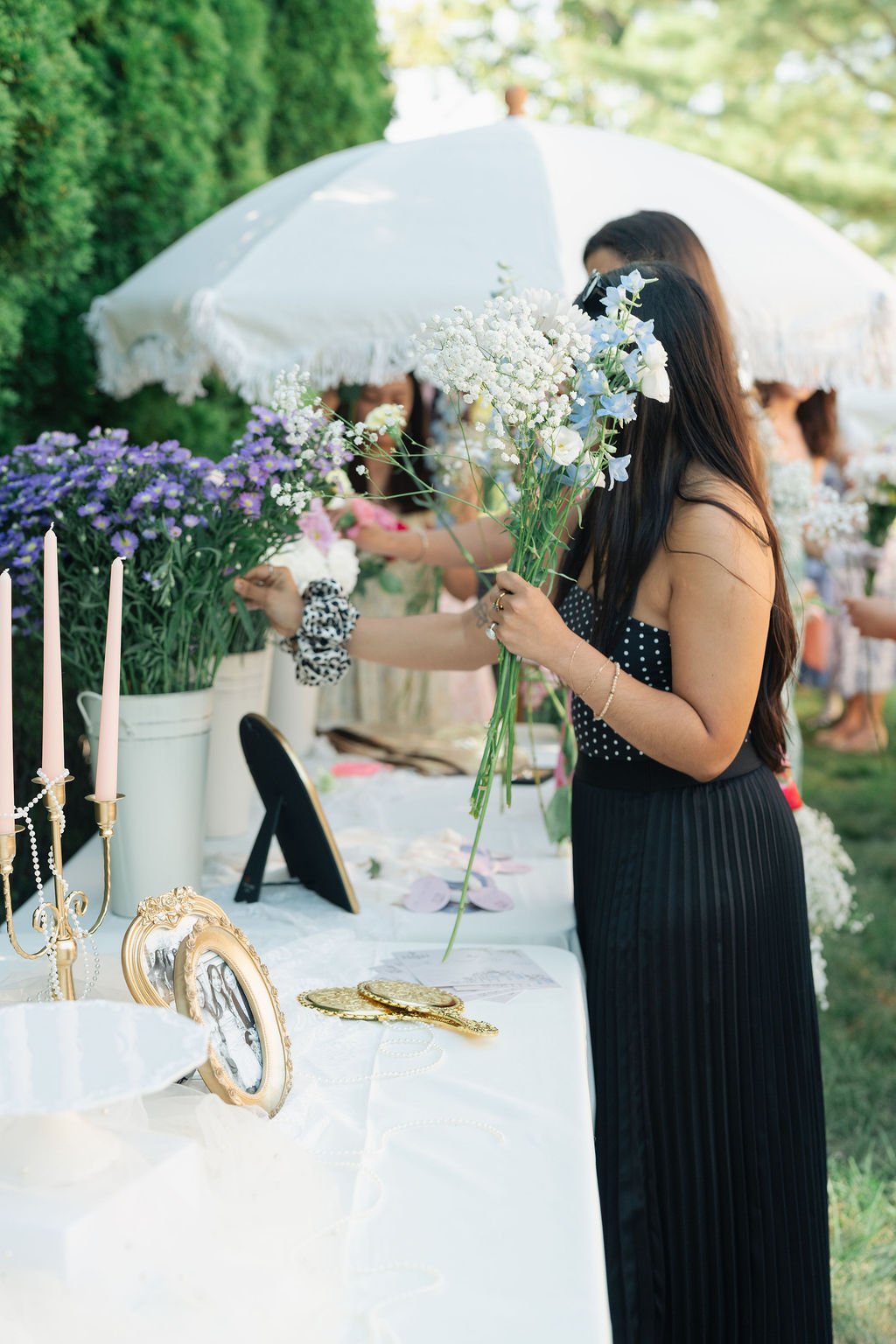 Woman in a black polka dot dress holding flowers at an outdoor event with other women and floral displays.