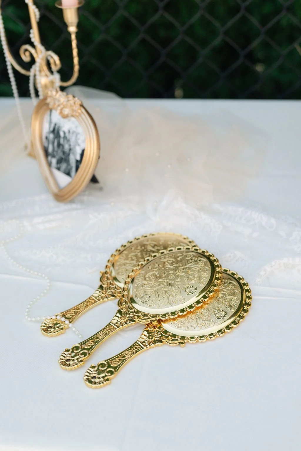 Three ornamental golden mirrors with intricate engraving, placed on a white fabric surface, with a white pearl necklace and a gold decorative frame in the background.
