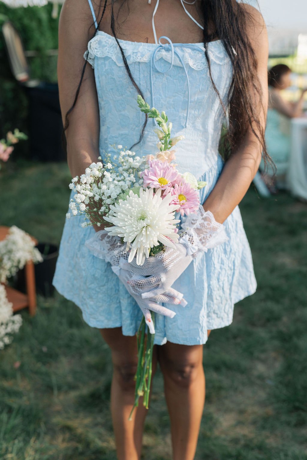 A woman is holding a bouquet of pink and white flowers, wearing a light blue dress with lace details and gloves, standing outdoors on a grass lawn.
