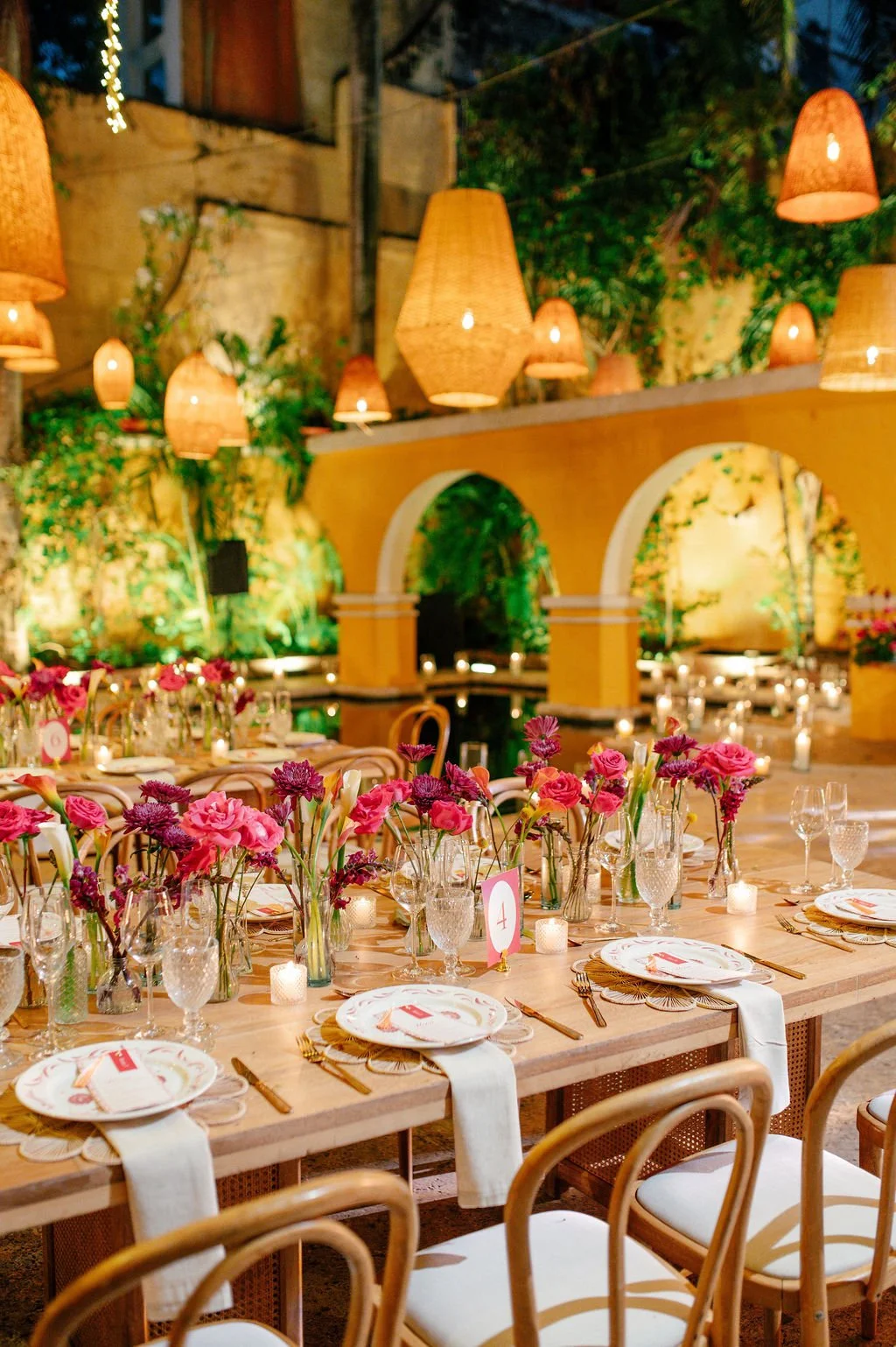 A beautifully decorated indoor dining area with long wooden tables set for a celebration. The tables are adorned with pink and purple flowers in glass vases, candles, and plates with gold utensils. The ambiance is warm with hanging wicker lamps and lush green foliage in the background.