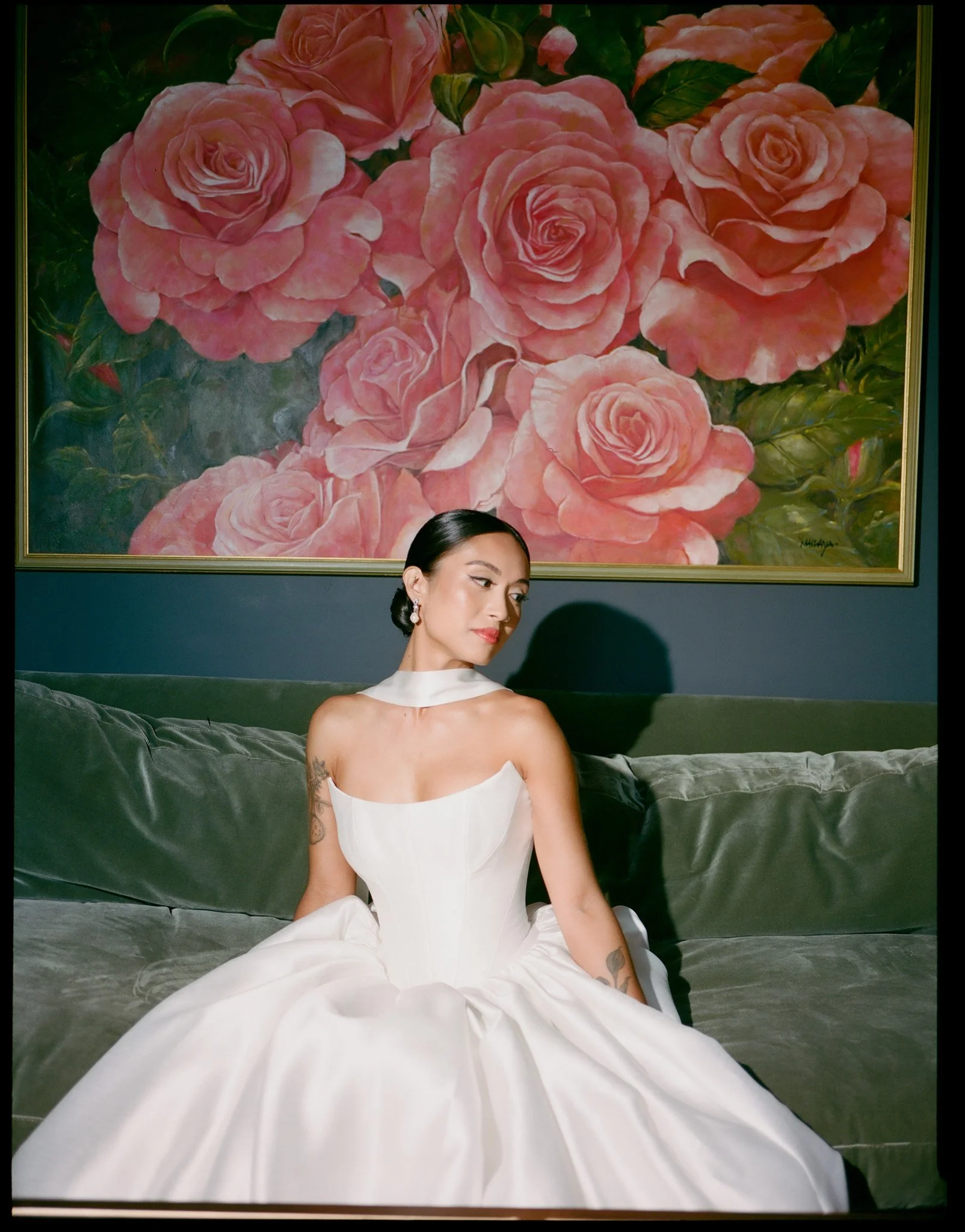 A woman in a white dress sitting on a sofa in front of a large painting of pink roses.