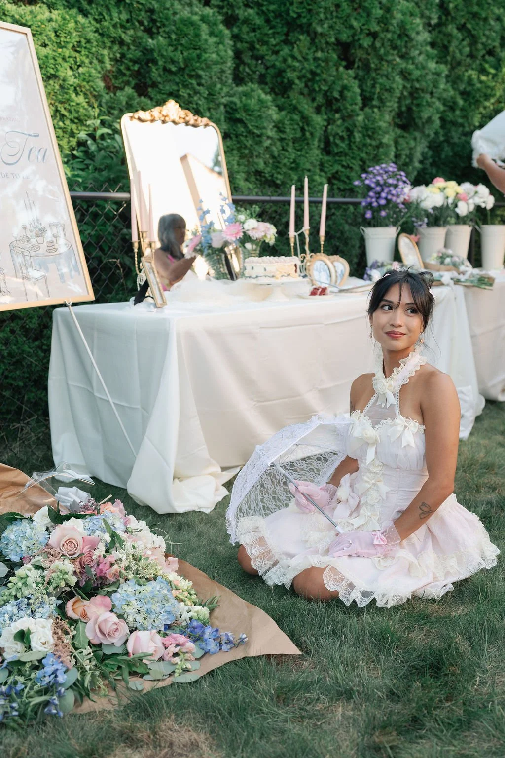 A woman in a vintage-style white dress with lace and bows, sitting on the grass holding a lace parasol, at an outdoor garden tea party with floral arrangements and decorated table.