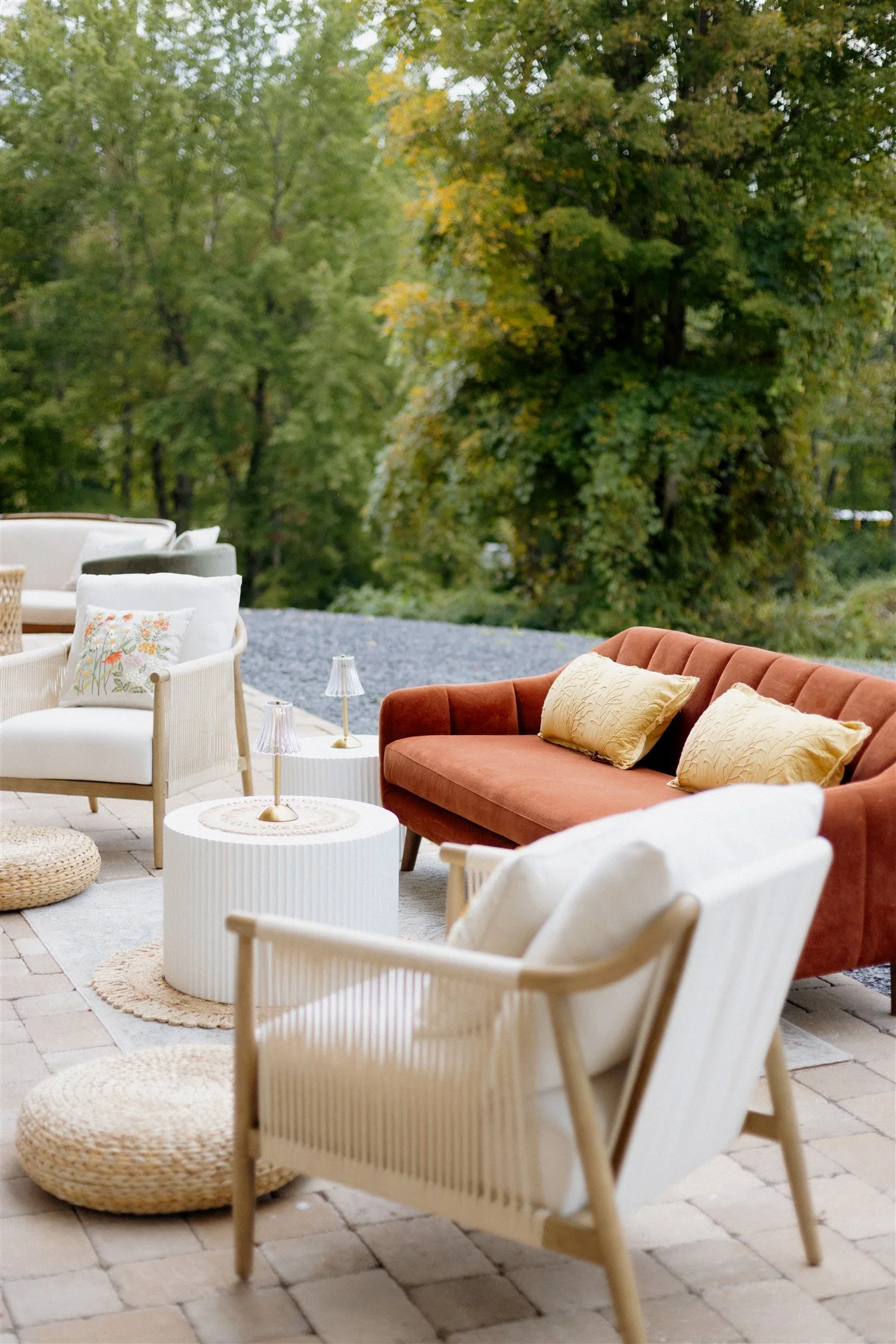 Outdoor patio with white wicker chairs, a rust-colored sofa with yellow pillows, and small tables with lamps, set against a backdrop of green trees.