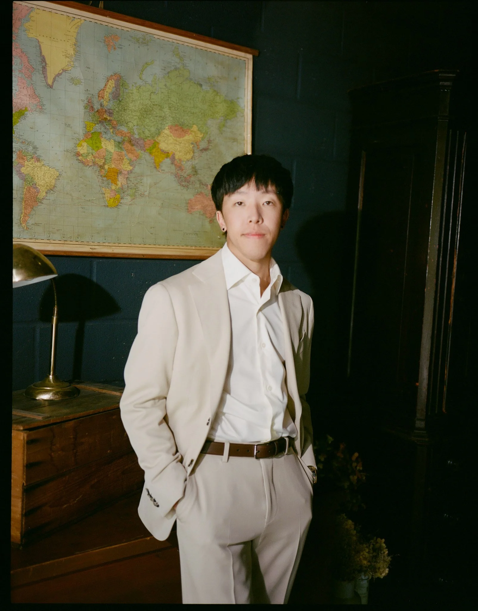 A man wearing a cream-colored suit and white shirt standing indoors in front of a world map. There is a dark cabinet to his right and a wooden dresser with a brass lamp in front of the map.