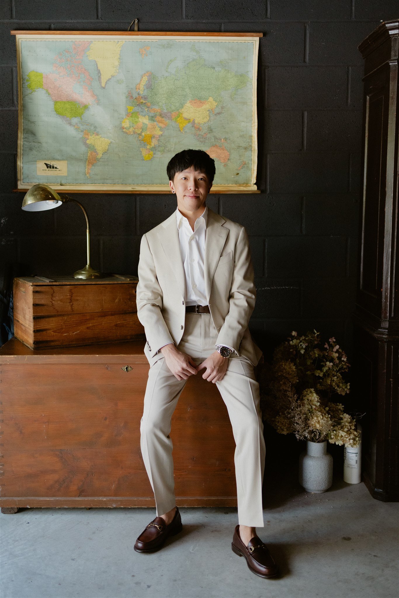 A young man in a white suit sits with legs apart in a room with dark brick walls. Behind him is a large world map and a desk with a brass desk lamp. To the right, there's a tall dark cabinet and a white vase with flowers on the floor.
