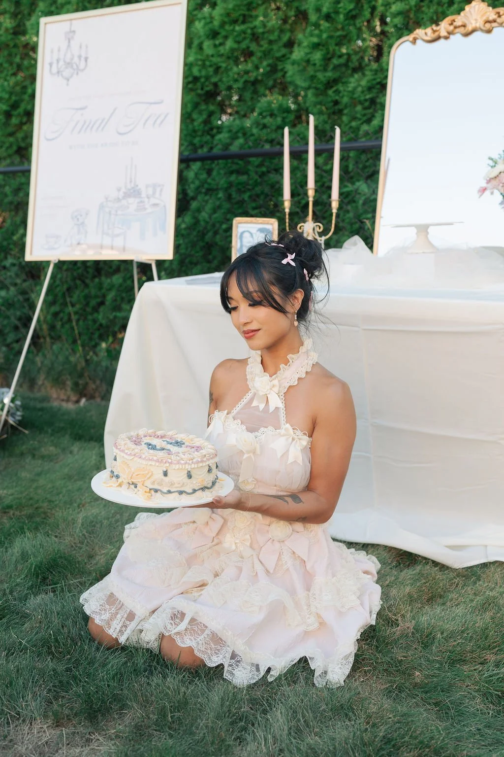 A woman in a lace dress holds a birthday cake at an outdoor celebration with a sign reading 'Final Fun' and a decorated table in the background.
