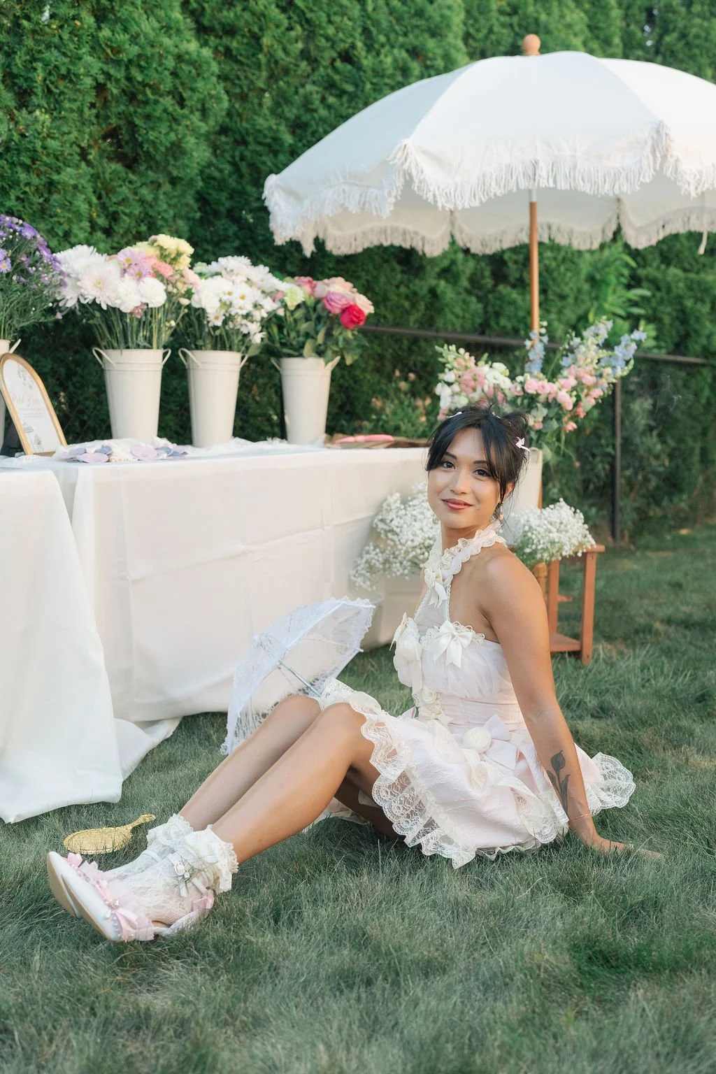 A woman in a white dress sitting on the grass at an outdoor event with flowers and a large white umbrella behind her.