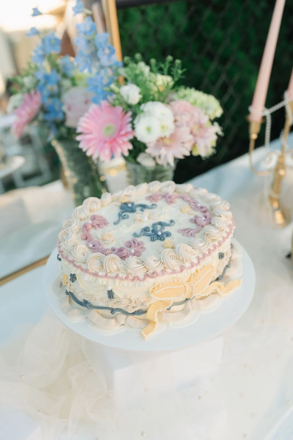 A decorated cake with floral and ribbon icing, placed on a white stand, with a bouquet of pink, white, and blue flowers in the background at an outdoor setting.