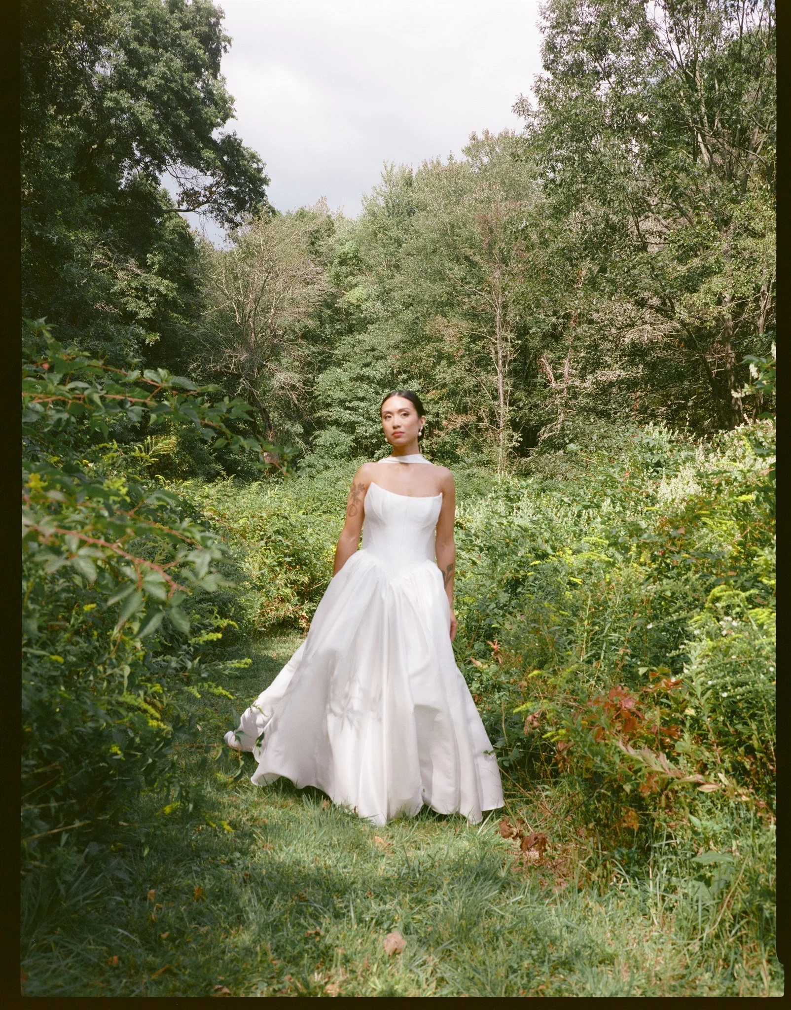 A woman with dark hair in an elegant white strapless gown standing in a lush green garden surrounded by dense leafy trees.