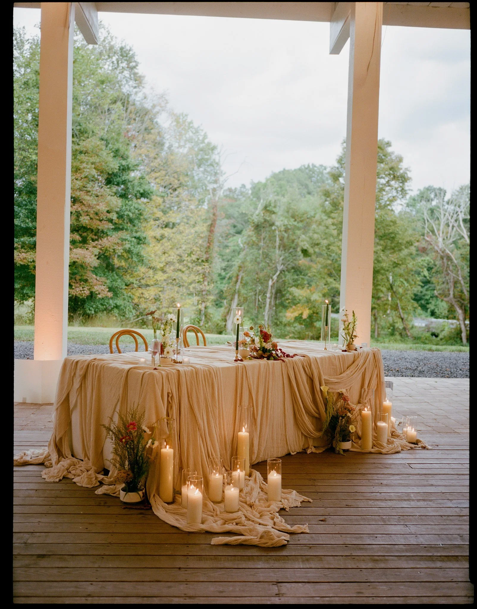 A decorated outdoor table with candles and floral arrangements, set on a wooden deck overlooking a lush green landscape with trees.