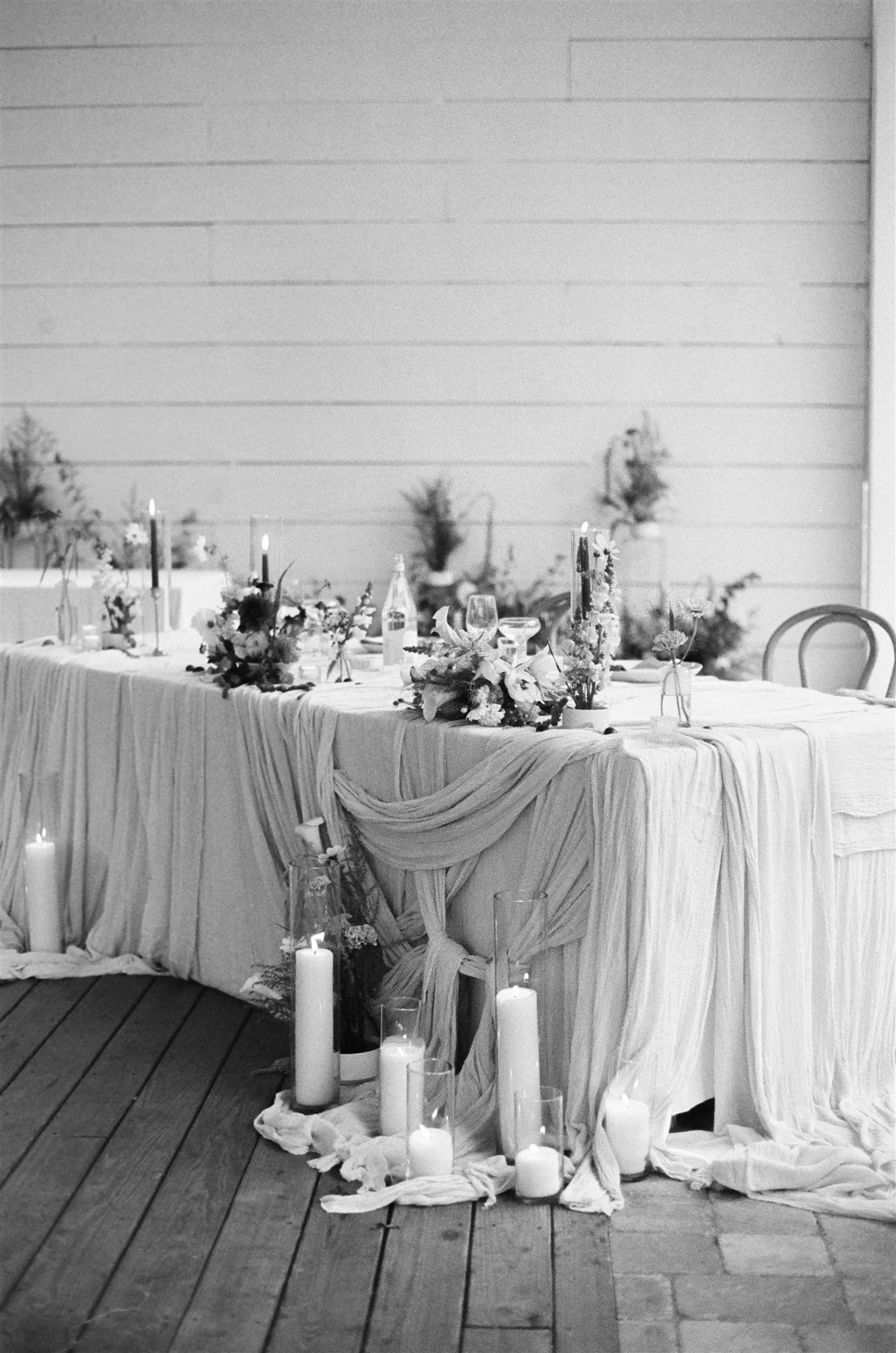 A long table decorated for a wedding or celebration with flowers, candles, and draped fabric, set on a wooden deck with candles on the floor nearby.