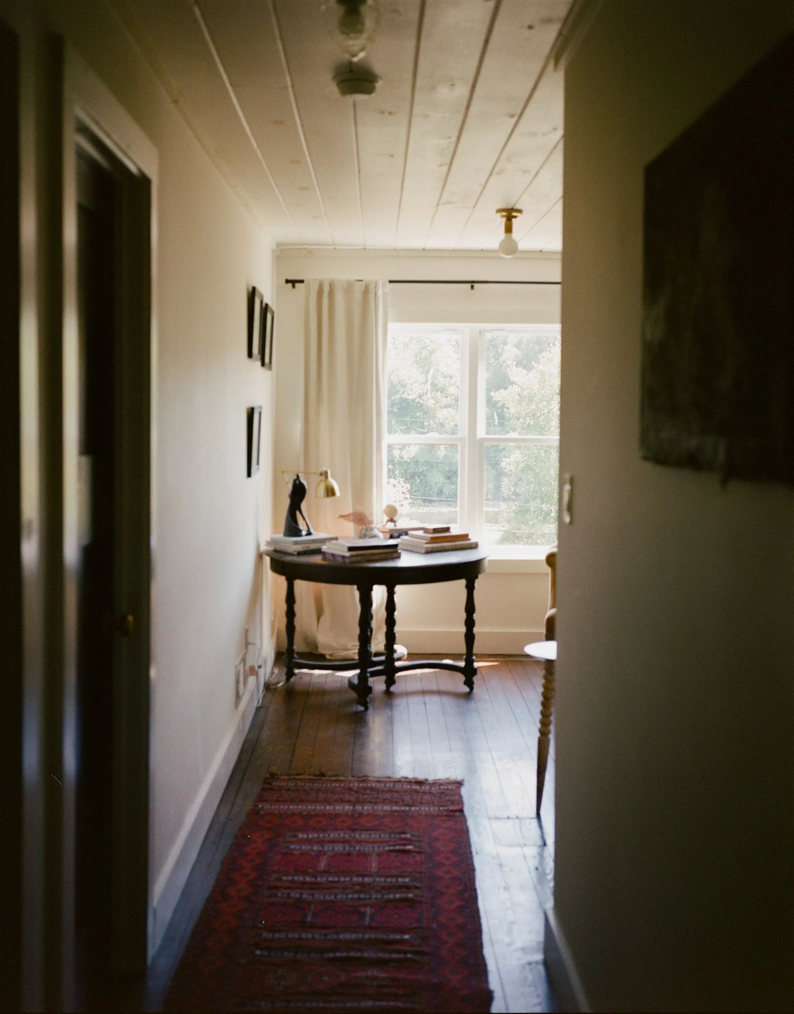 An indoor hallway with a wooden floor, a red patterned rug, a small round table with stacked books, a black sculpture, a lamp, and a window with white curtains letting in natural light.