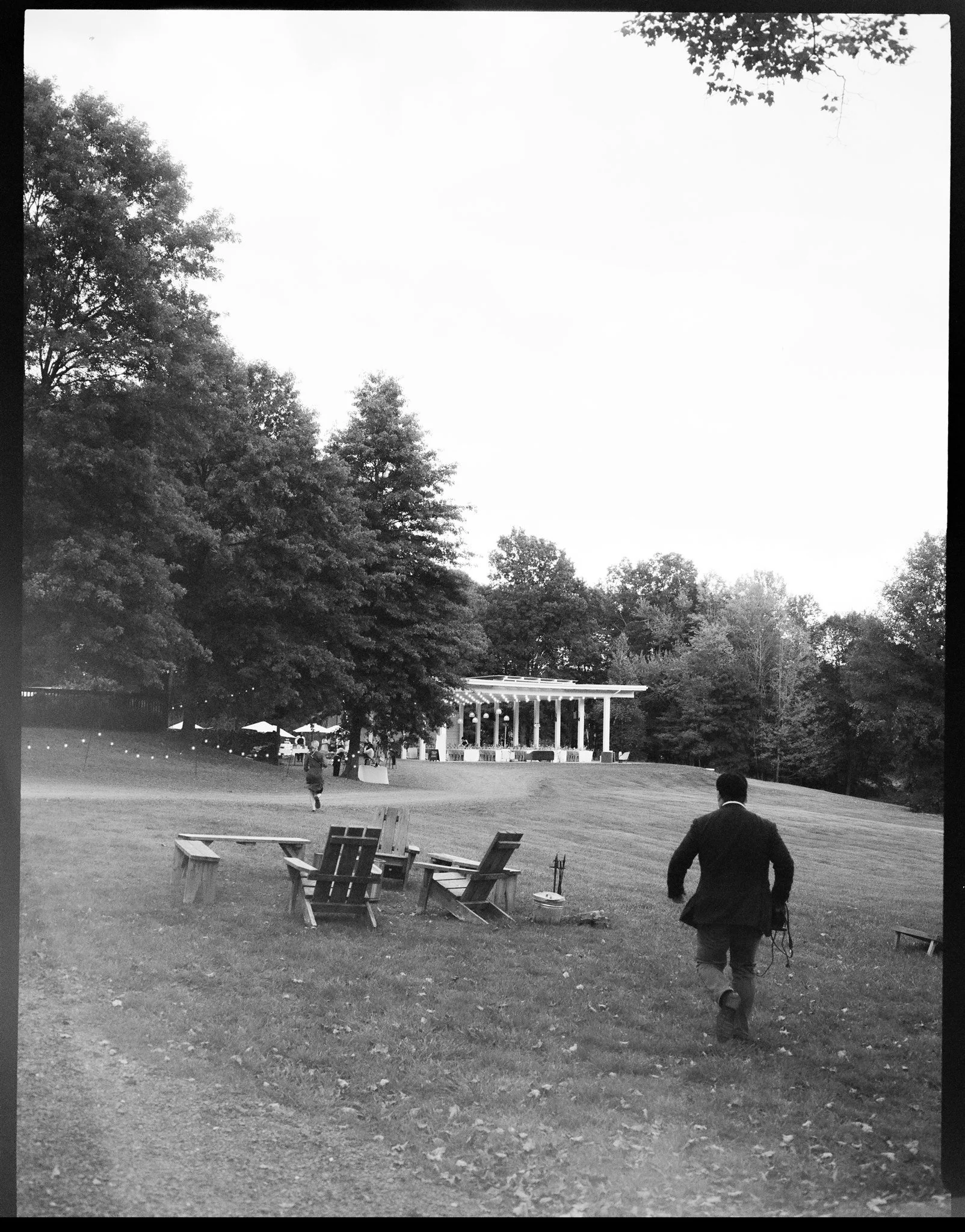 Black and white photo of a park with trees, benches, and a bandstand. One person in a suit is walking away from the camera, holding a bag.