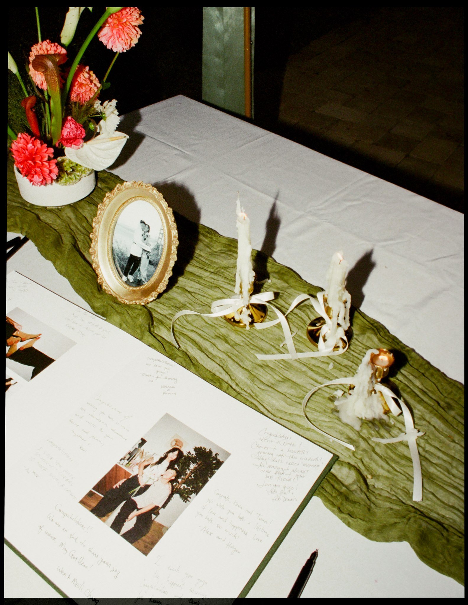 A table with a green wooden surface holding a white floral arrangement, an older picture frame with a black-and-white photo of children, three lit candles with decorative white ribbons, and handwritten notes on white paper. The background appears to 